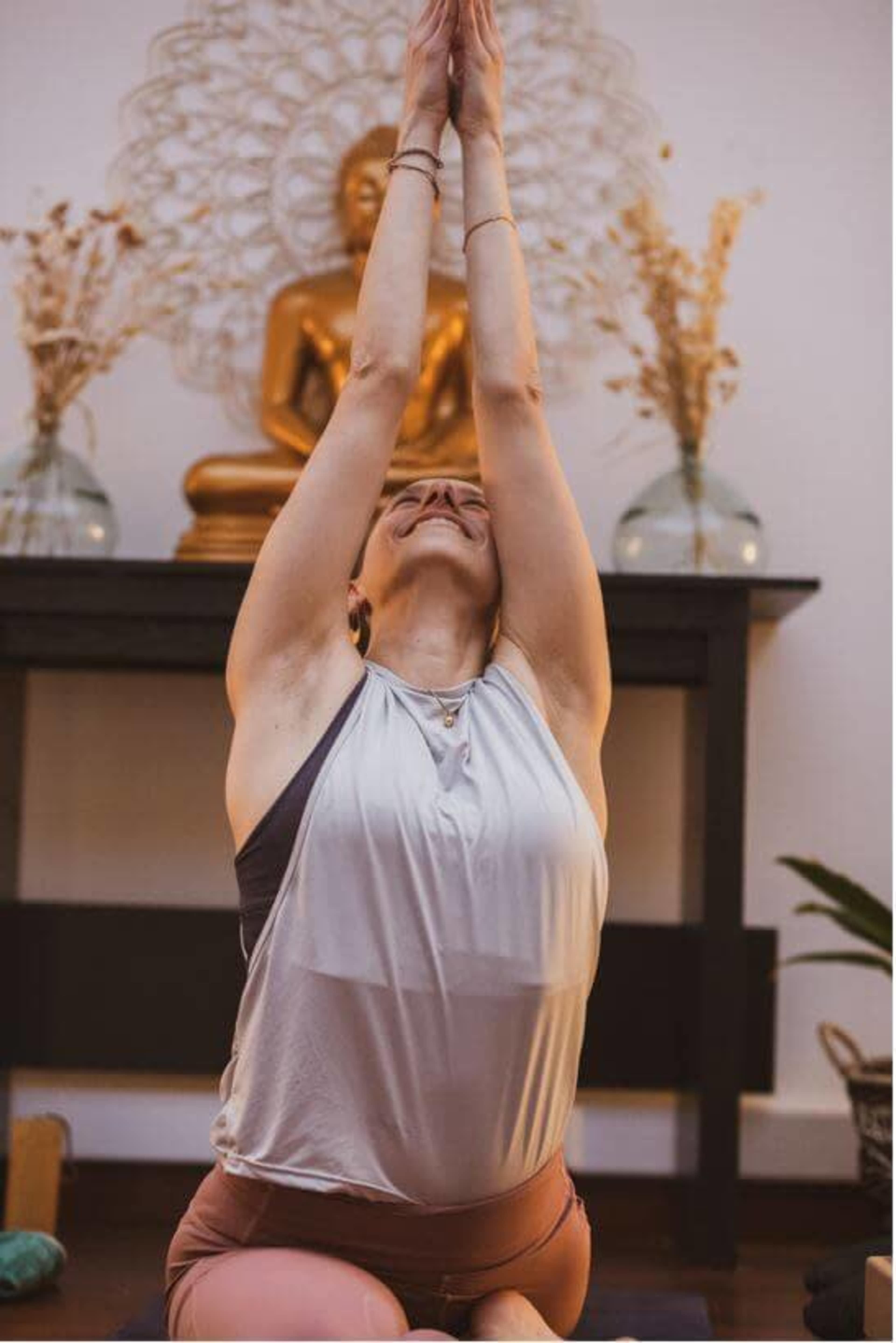 A person performs a yoga pose with hands clasped overhead in front of a seated Buddha statue and decorative elements.