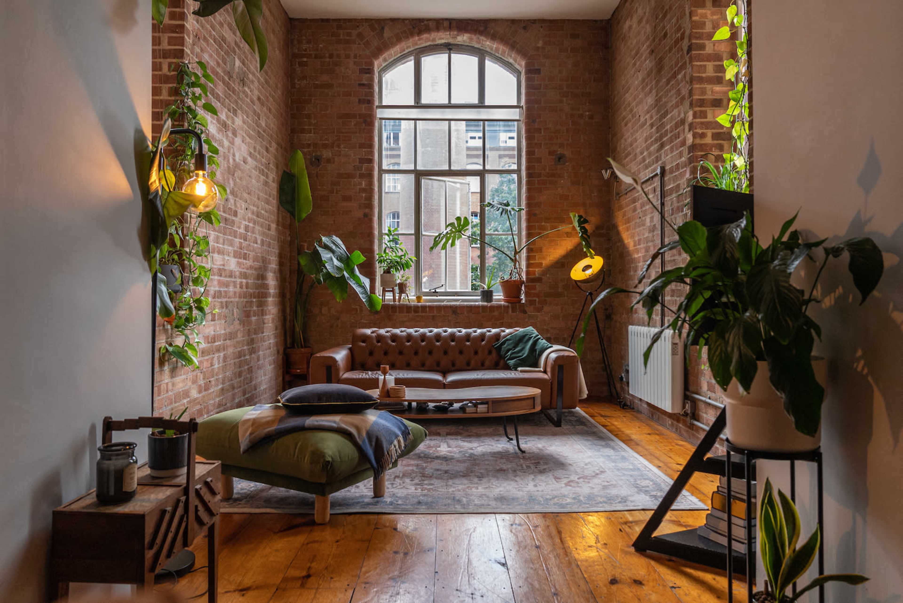 A cozy living room featuring a brown leather sofa, a patterned rug, wooden flooring, and large windows with plants positioned throughout the space.