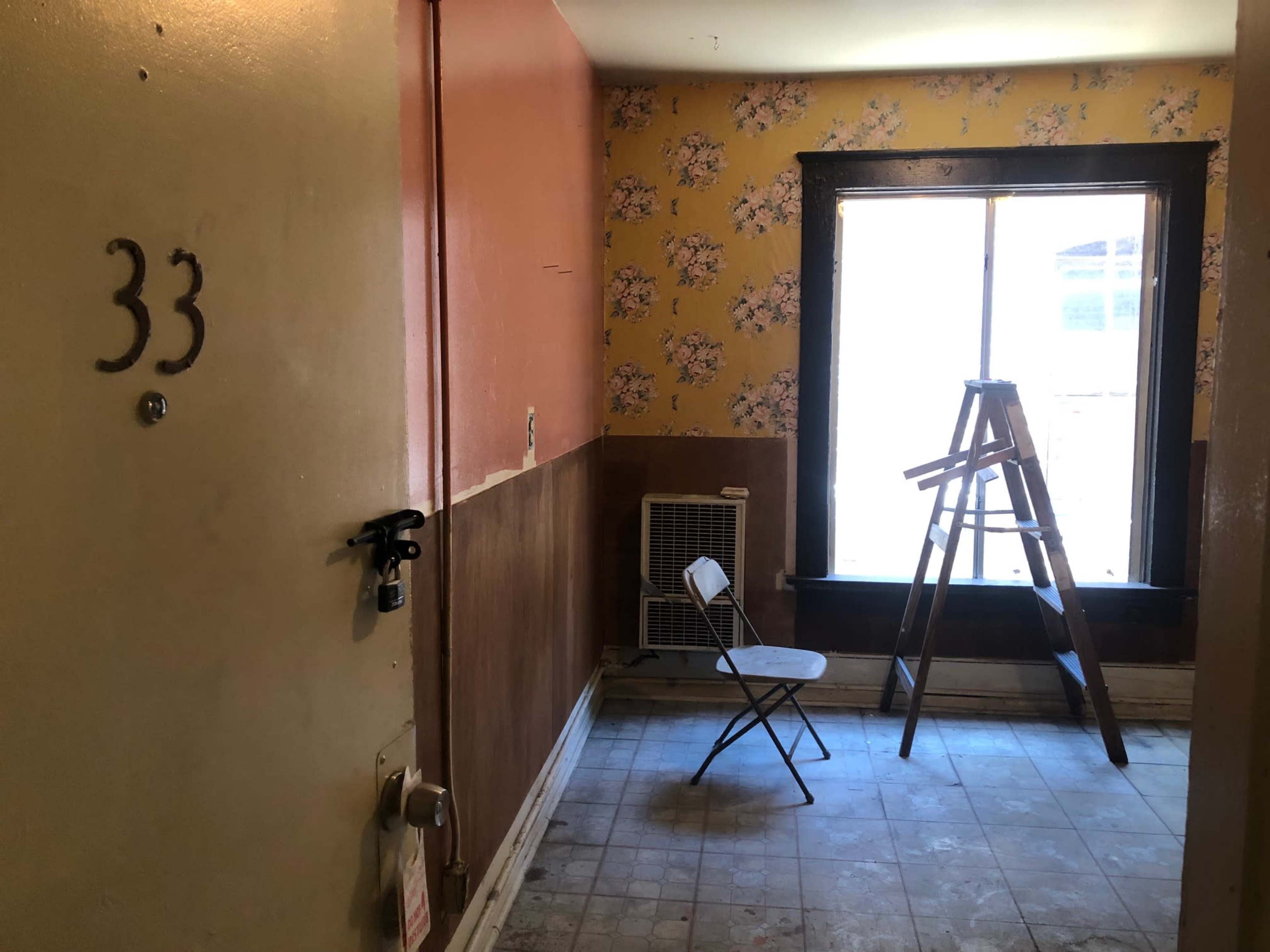 The image shows an unfinished room with a partially painted wall, a ladder, a chair, and a window.