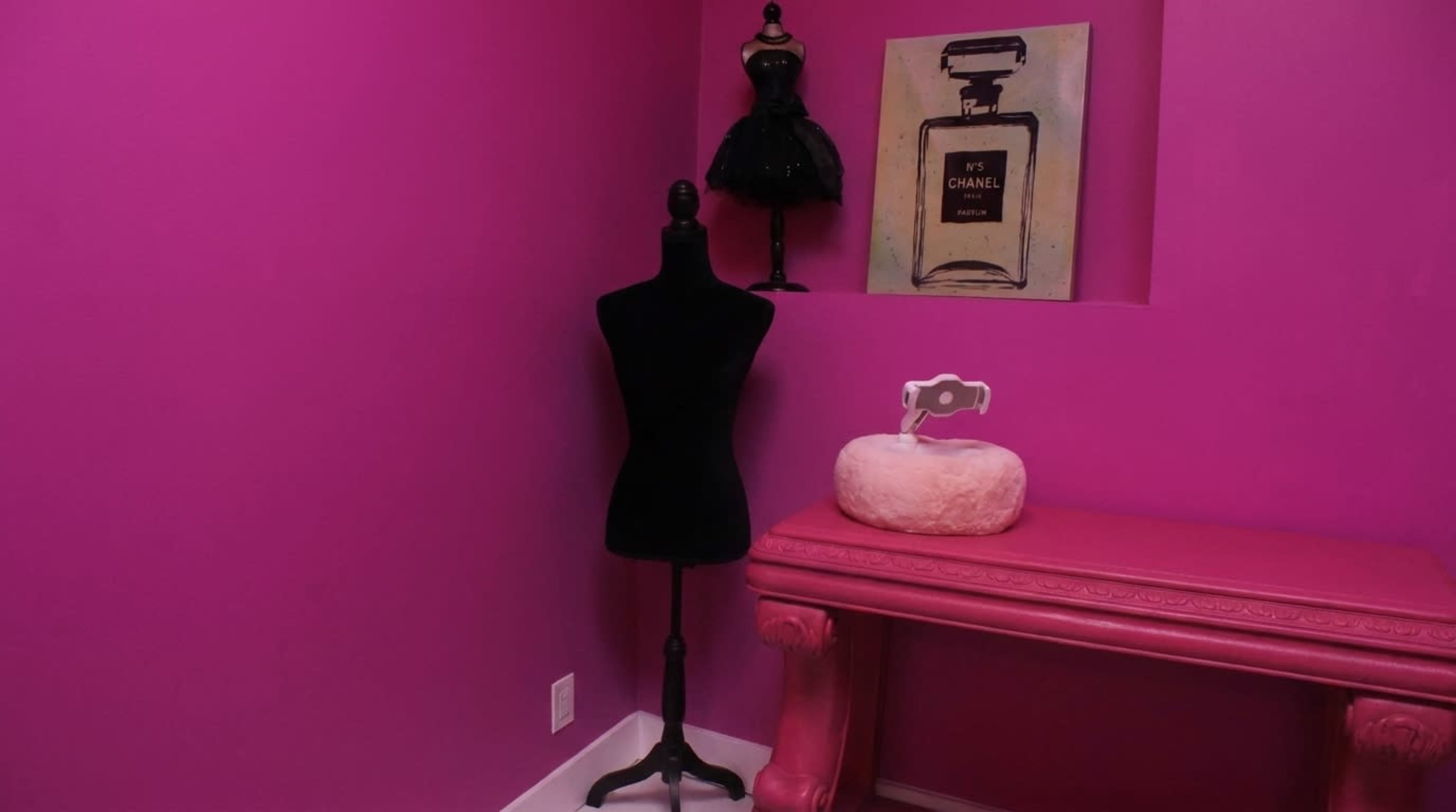 The image features a pink room with a black dress form, a pink table, and a framed artwork of a Chanel perfume bottle on the wall.