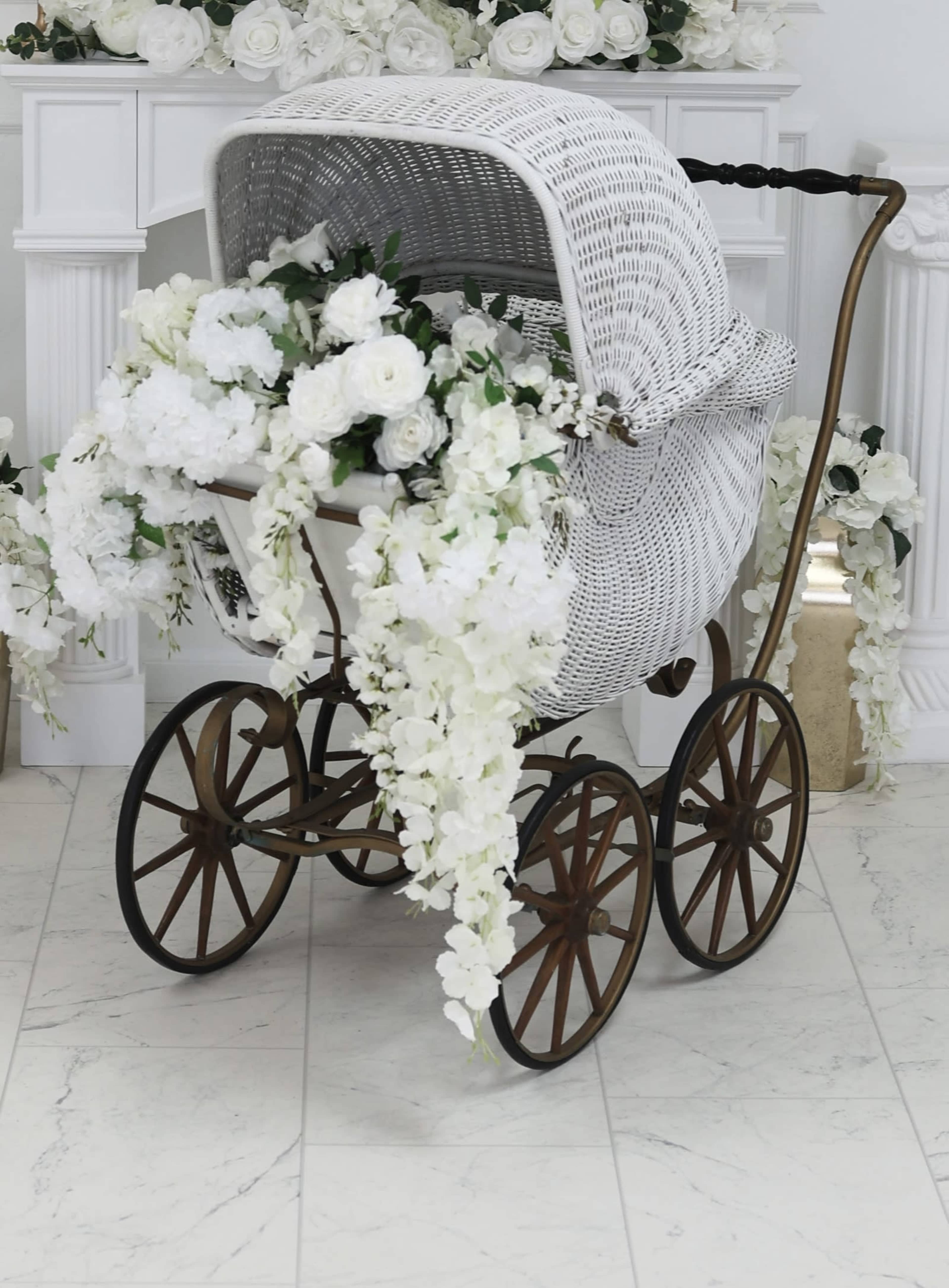 A vintage white wicker baby carriage adorned with cascading white flowers sits on a marble floor, surrounded by floral decorations.