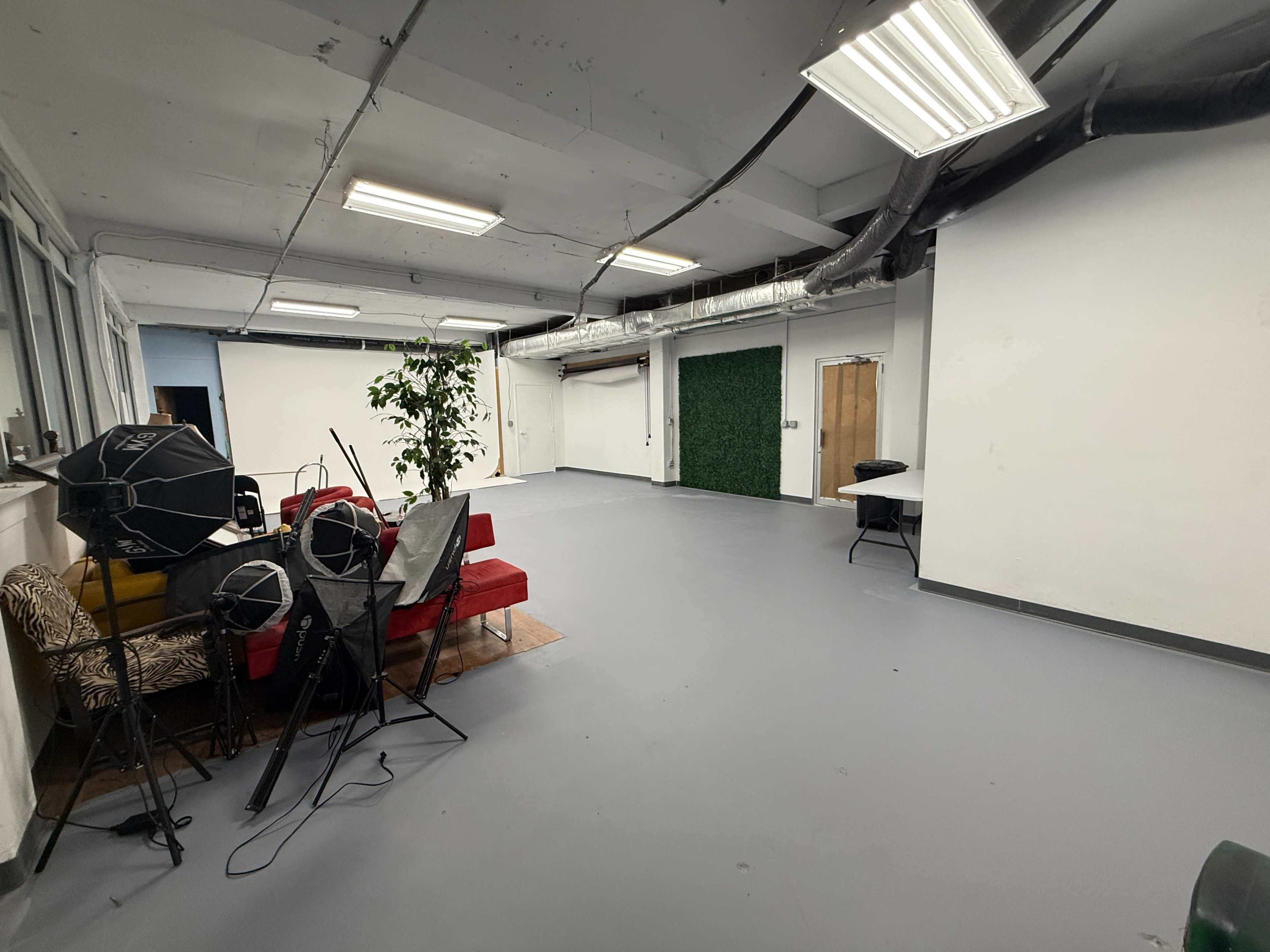 The image shows a spacious, empty room with gray flooring, a few chairs, a couch, and photography equipment scattered around.