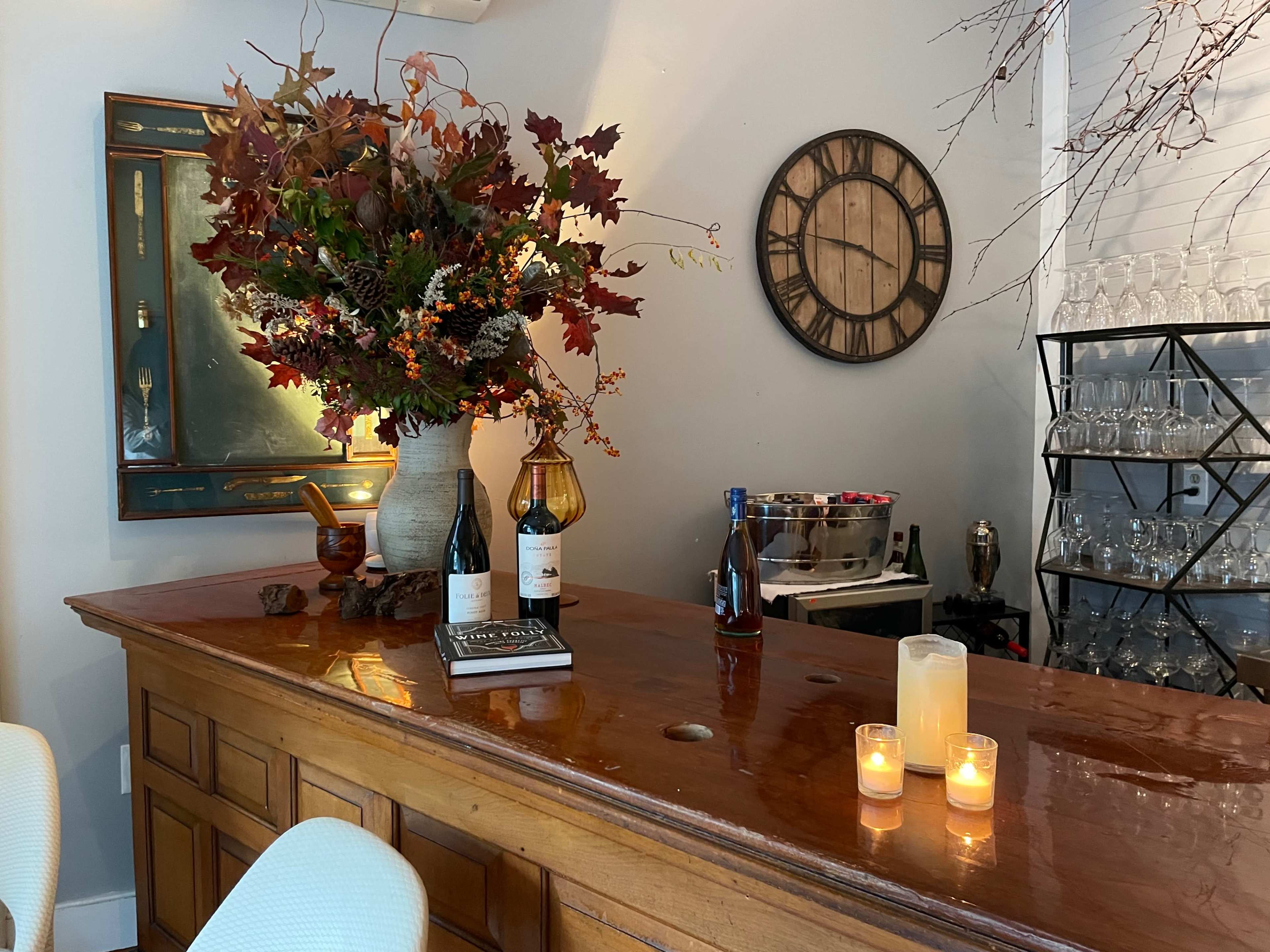 A wooden bar displays a floral arrangement, wine bottles, books, and lit candles, with a clock and glassware in the background.