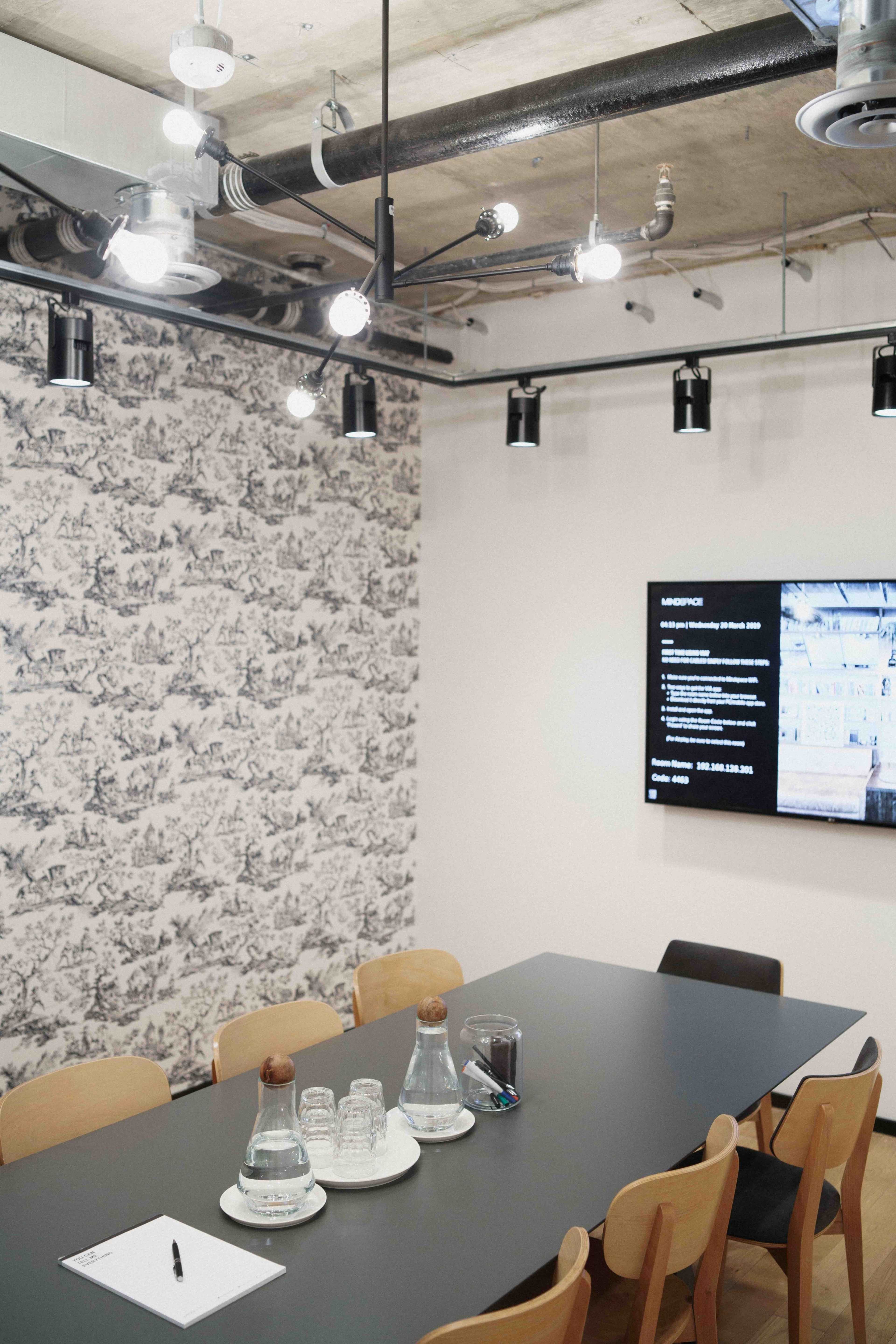 A modern conference room features a long dark table, wooden chairs, a projector screen displaying information, and decorative wallpaper.
