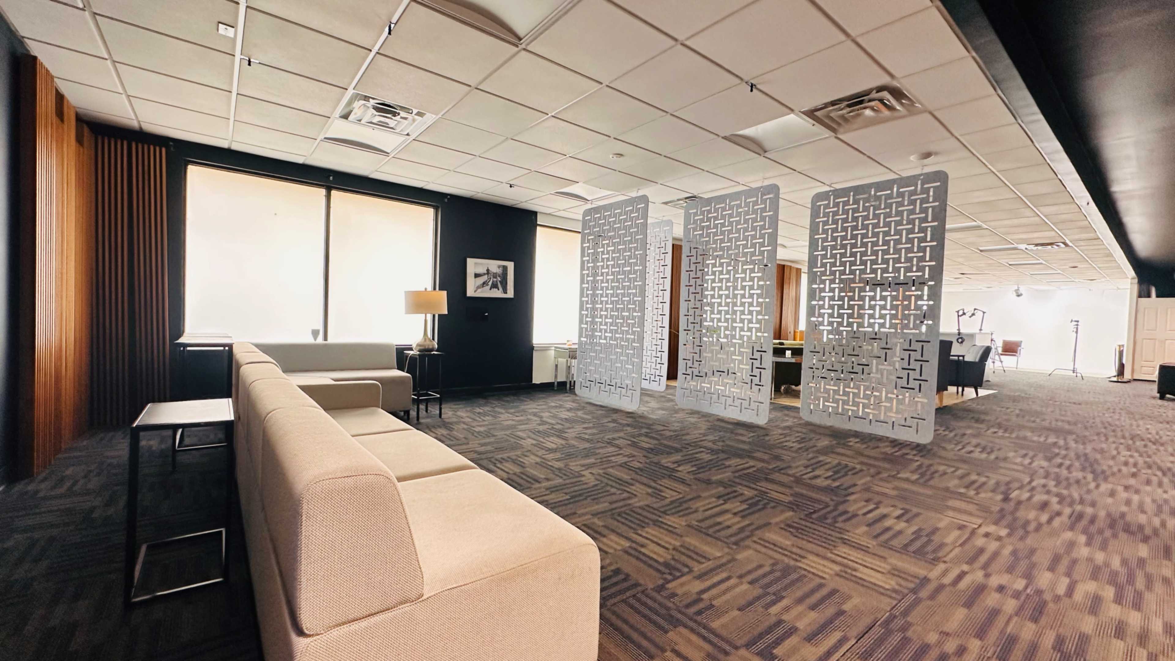 The image shows a modern office interior featuring a light-colored sofa, a lamp, and decorative partition screens, with large windows providing natural light.