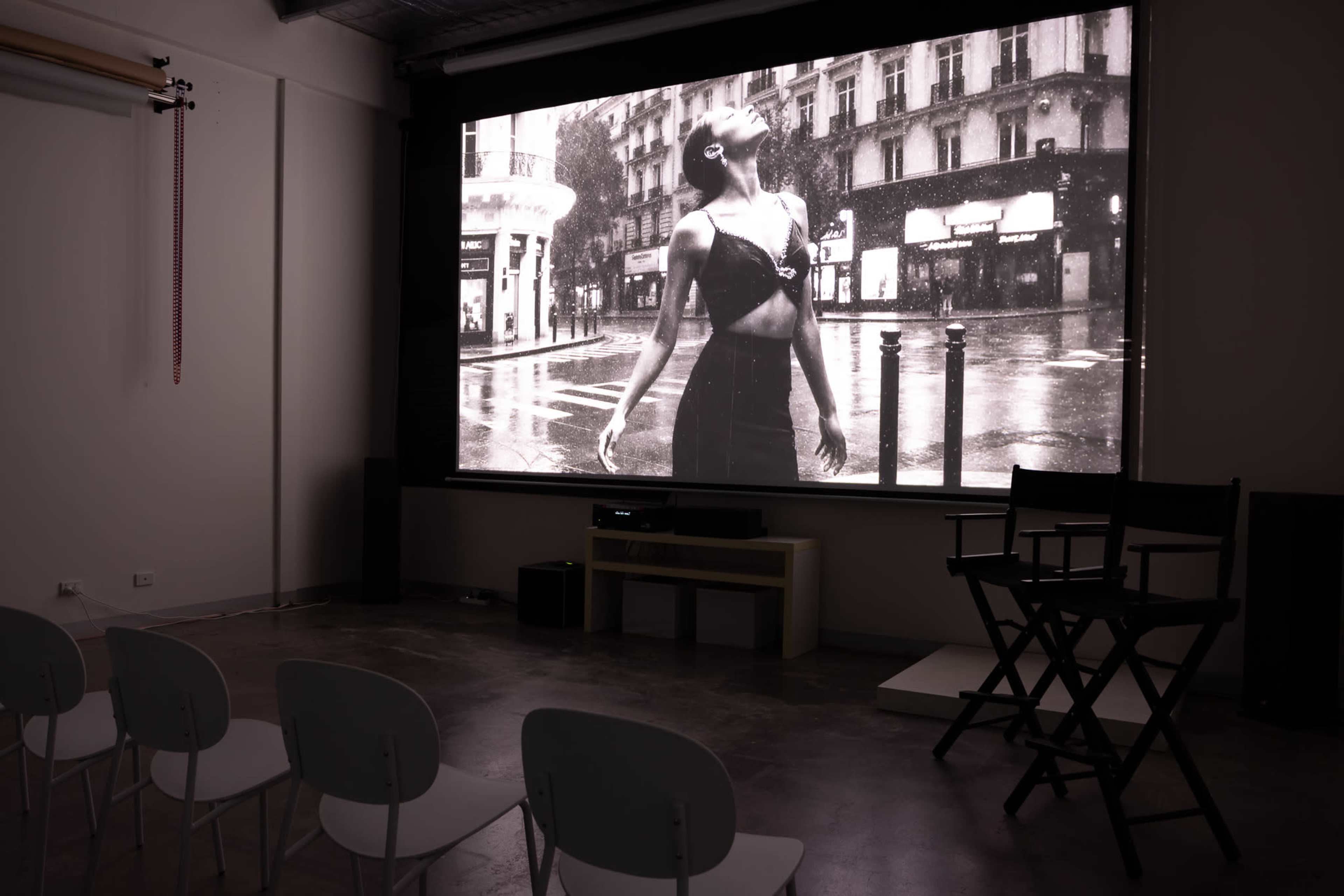 A large black-and-white photograph of a woman in a city rain scene is displayed on a screen in a room with empty chairs and a director's chair.
