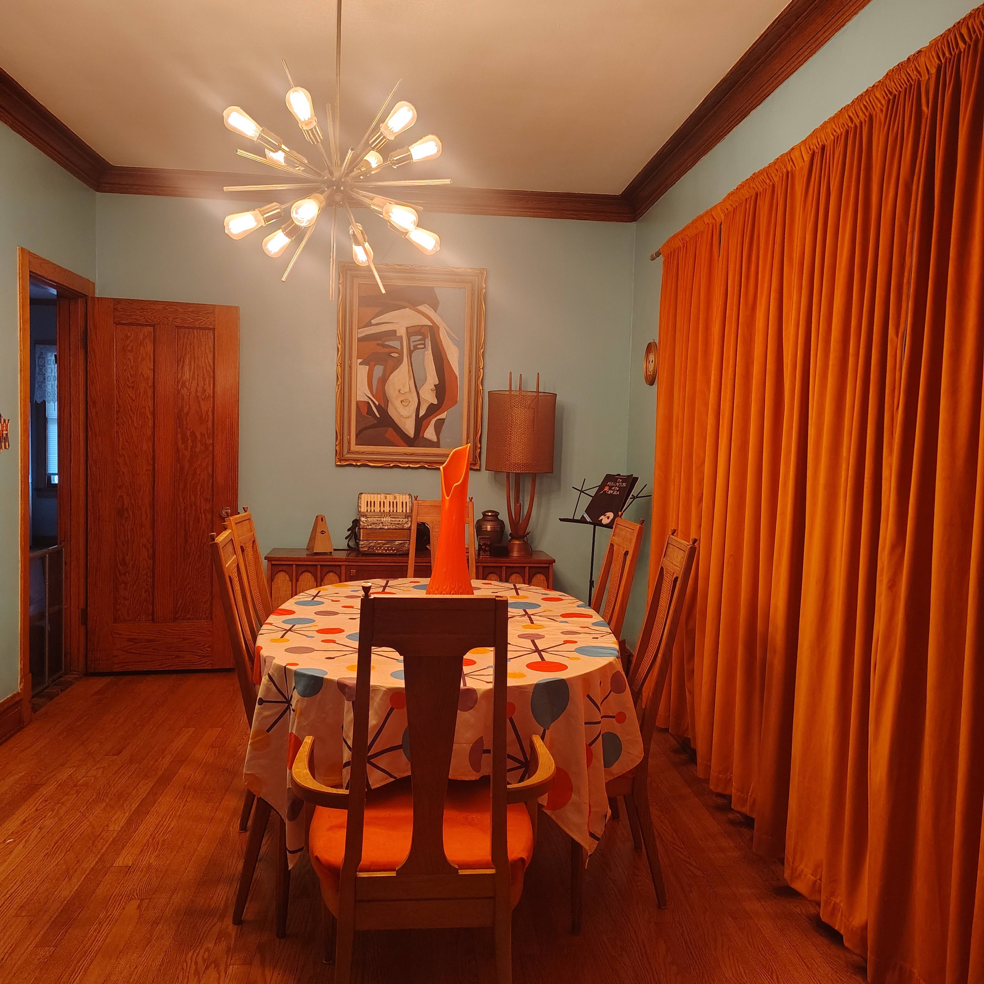 The image shows a dining room with a wooden table covered by a patterned tablecloth, surrounded by wooden chairs, and illuminated by a modern chandelier, with orange curtains and a painting on the wall.