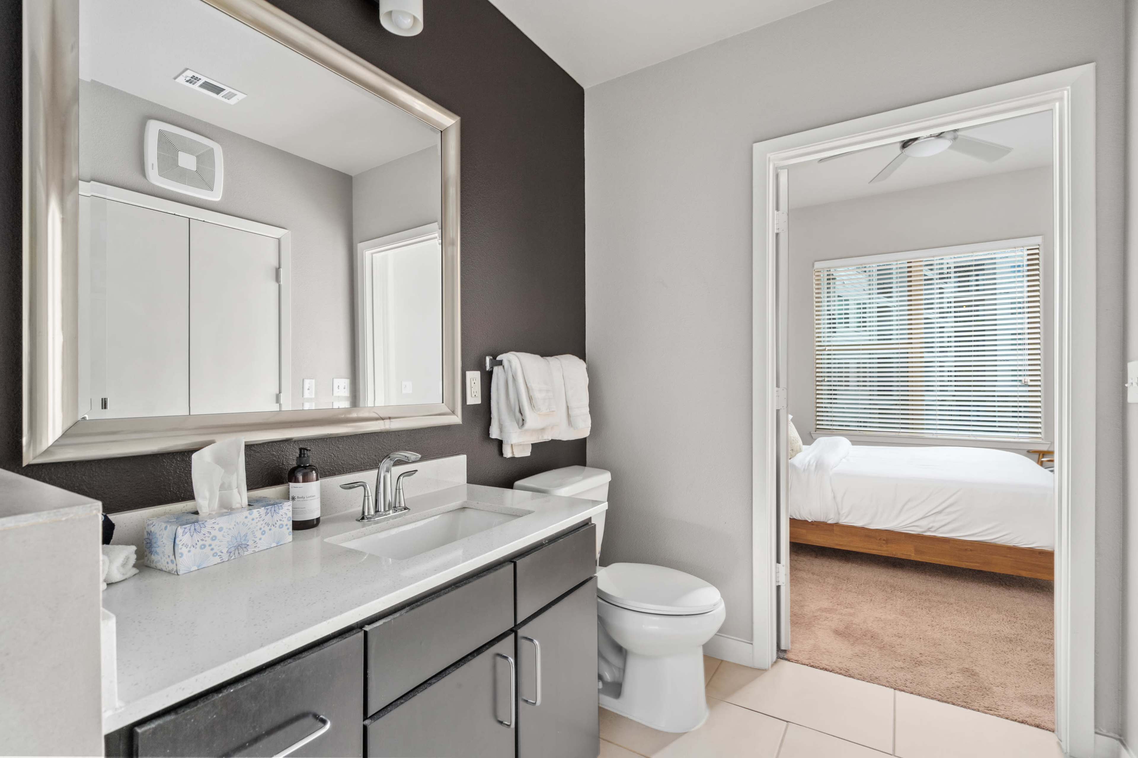A bathroom features a double sink vanity with a large mirror and a door leading to a bedroom with a bed and window.