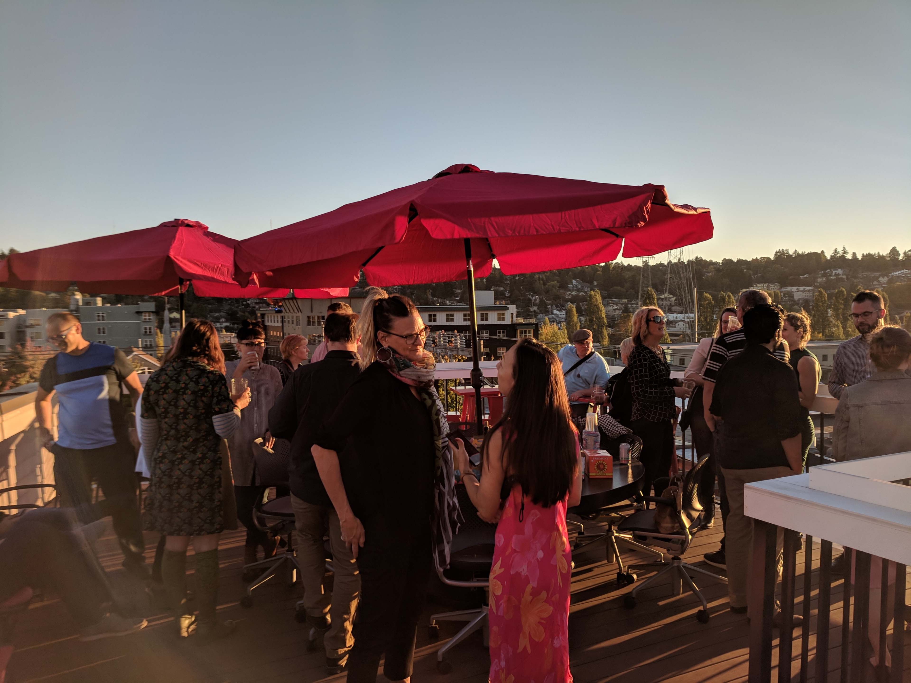 A group of people socializes under red umbrellas on a rooftop during sunset.