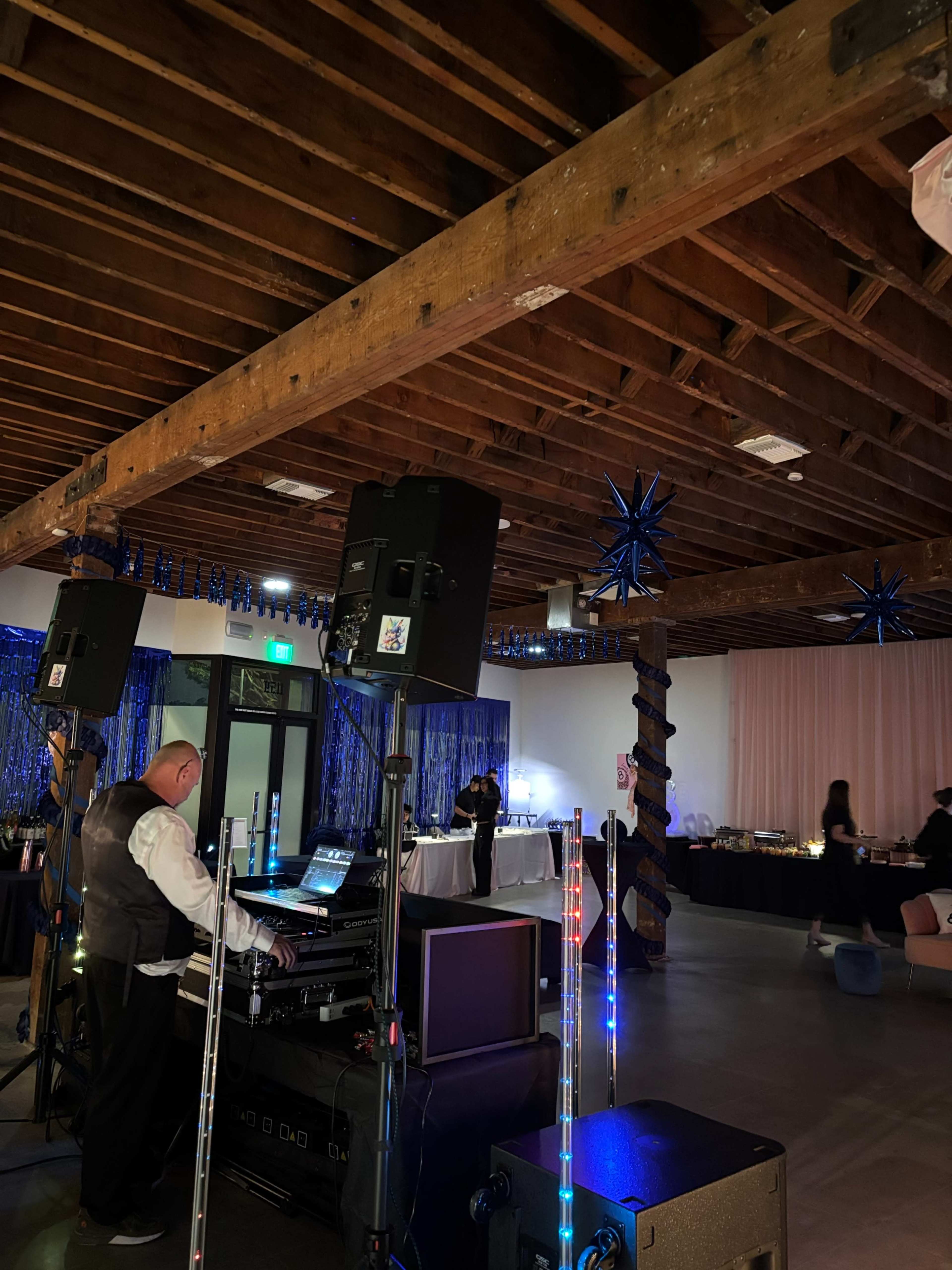 A DJ sets up equipment in a venue decorated with blue accents, while people mingle in the background near a food display.