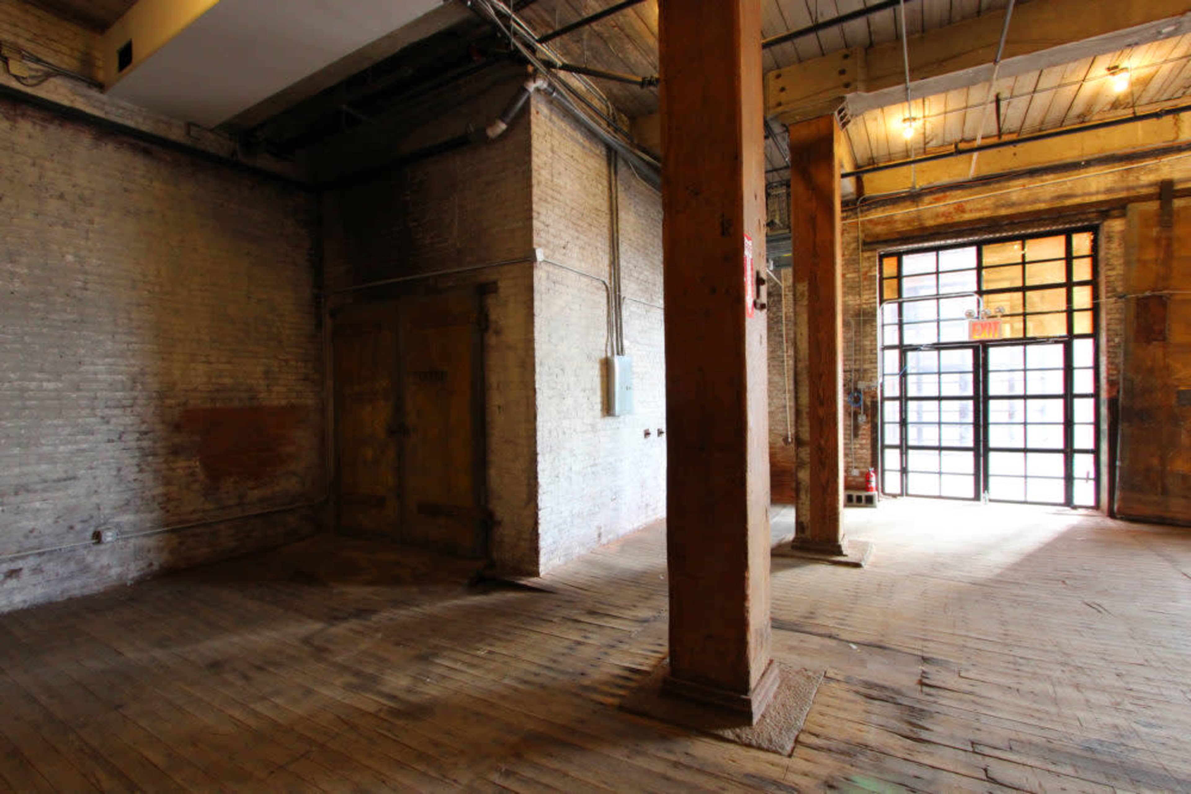 An empty room with brick walls, wooden floors, and several structural columns, leading to a large glass door.