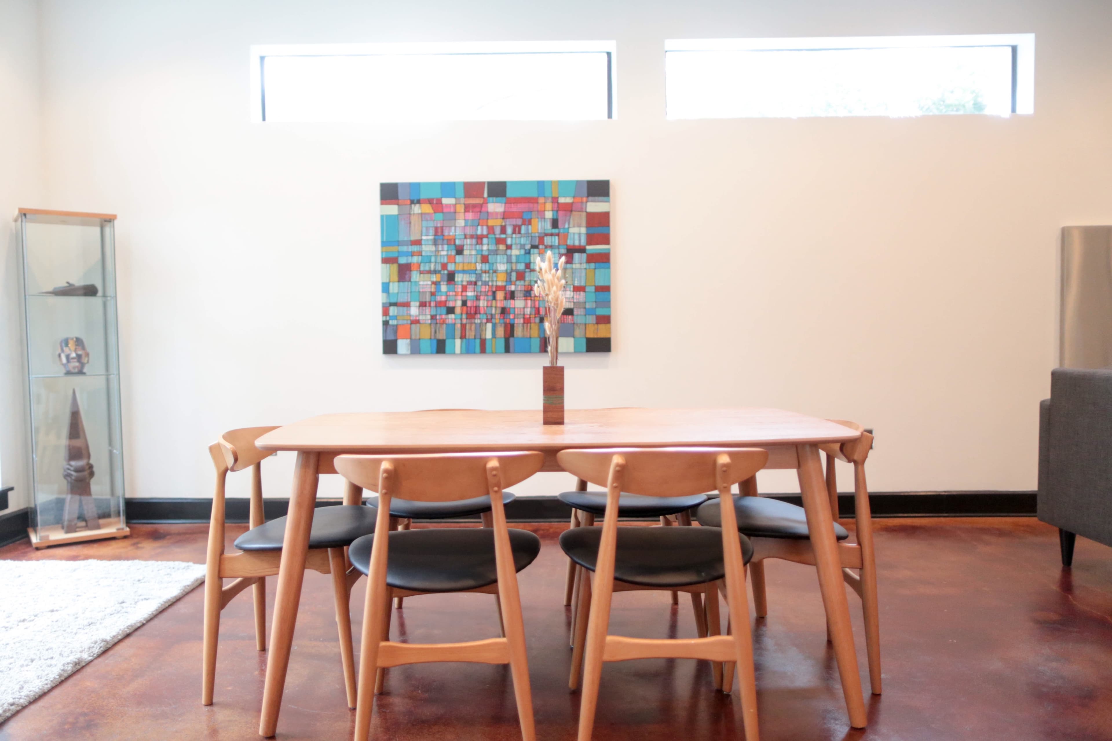 A wooden dining table with black chairs is set in a room featuring a colorful abstract painting and a display case.