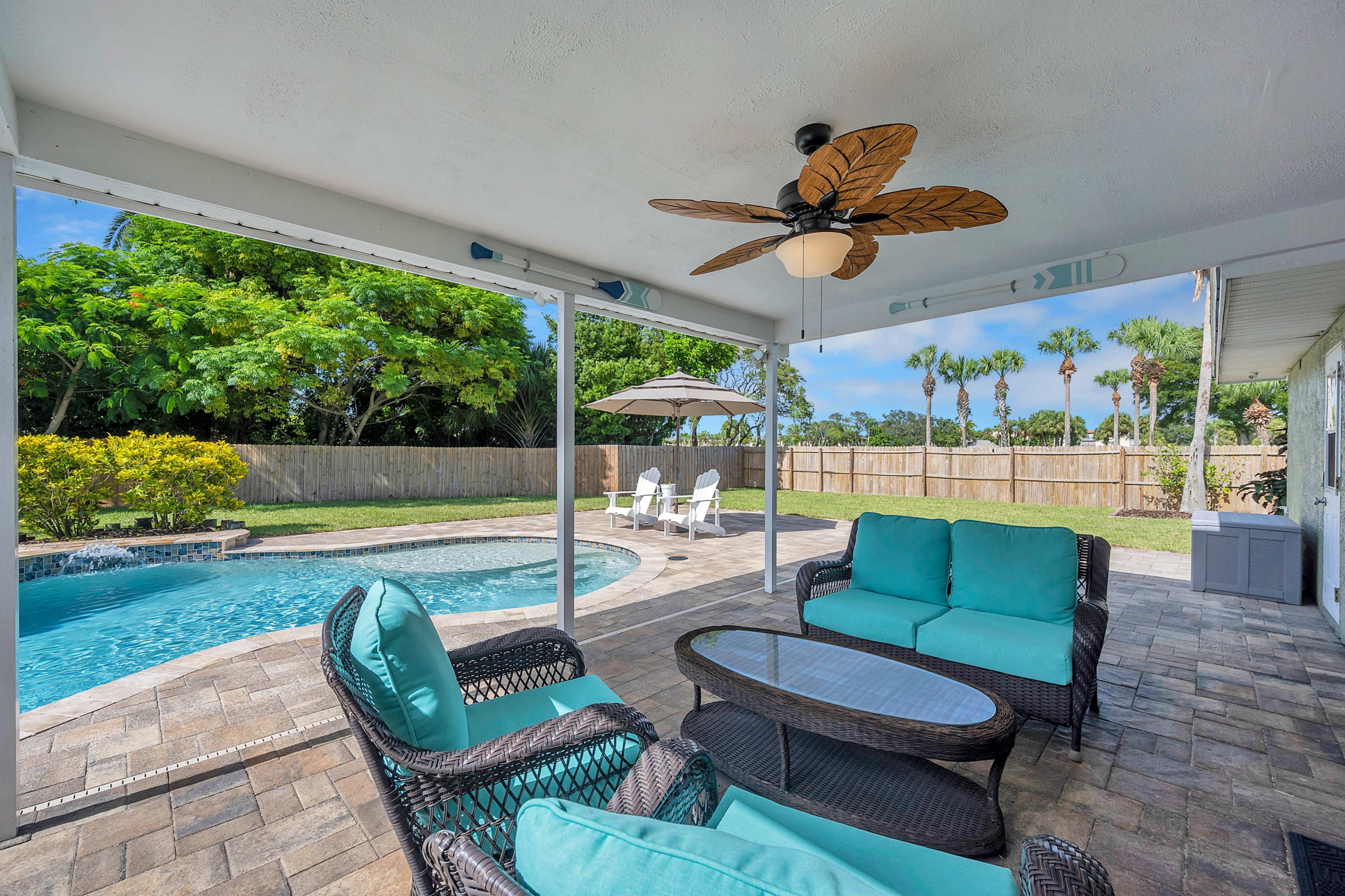 A covered patio area with wicker furniture and a ceiling fan overlooks a pool surrounded by a paver deck and tropical landscaping.