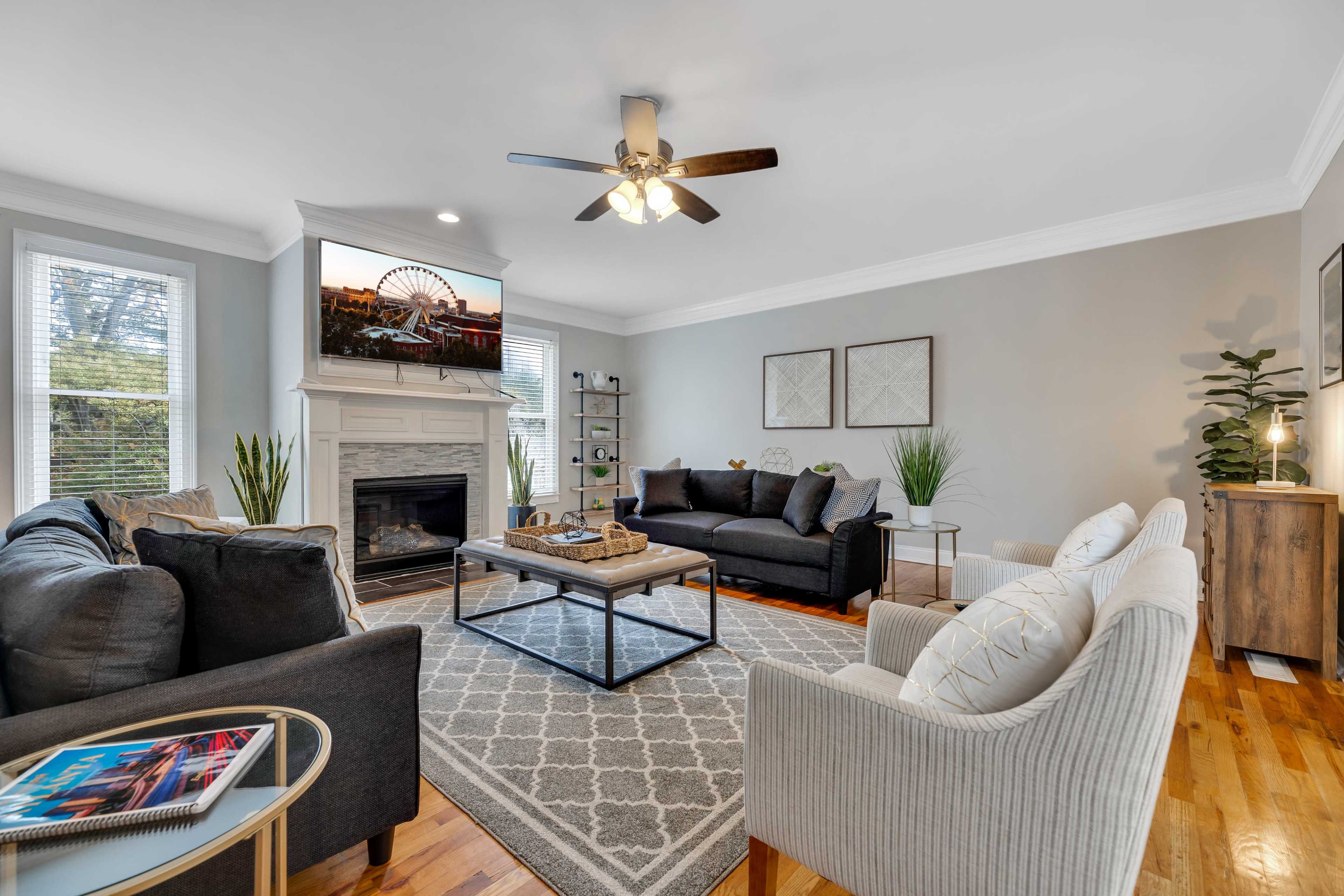 A modern living room with a fireplace, two sofas, a coffee table, and a television mounted above the mantel, surrounded by plants and decorative items.