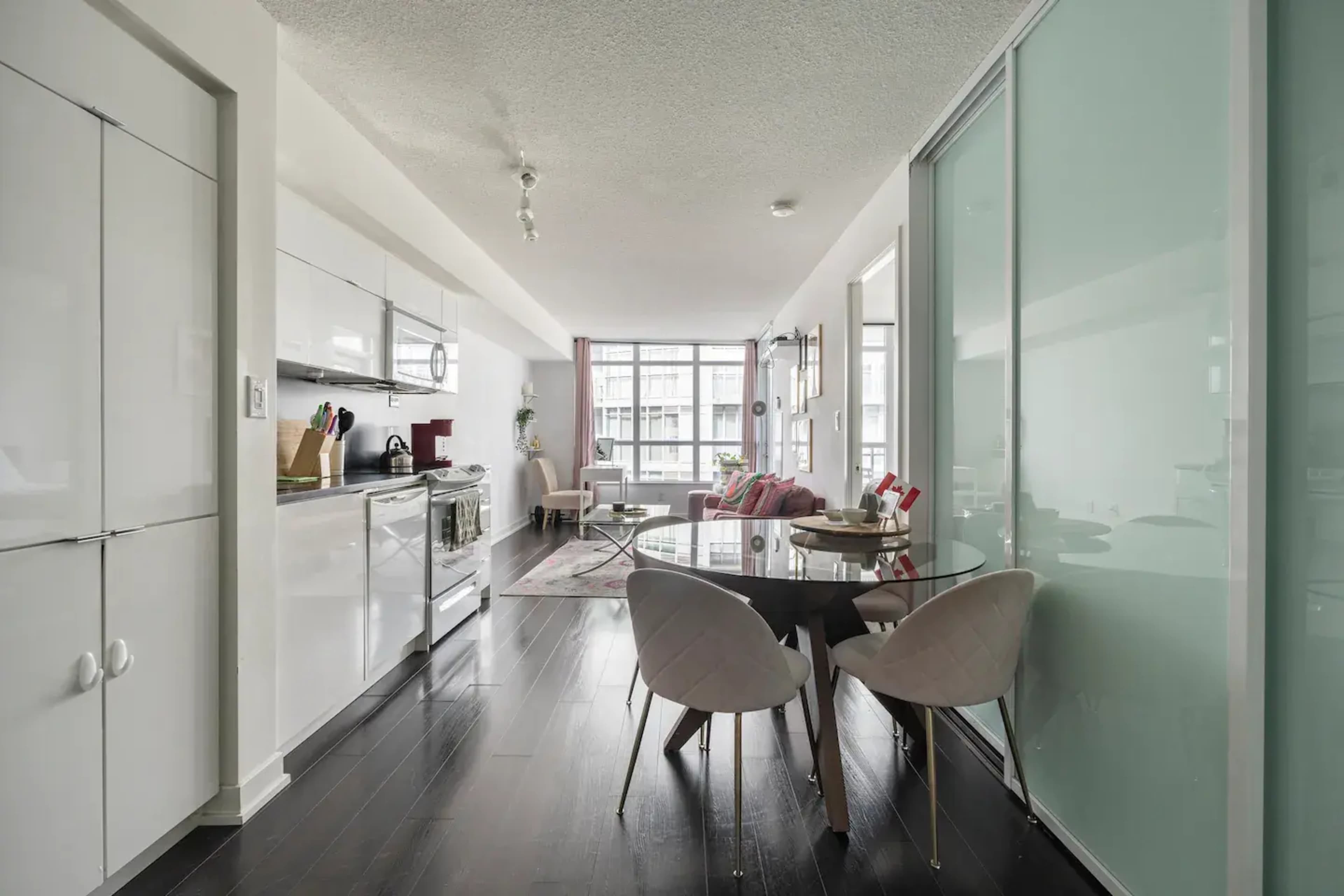 A modern kitchen and dining area features light cabinetry, a round table with chairs, and large windows allowing natural light.