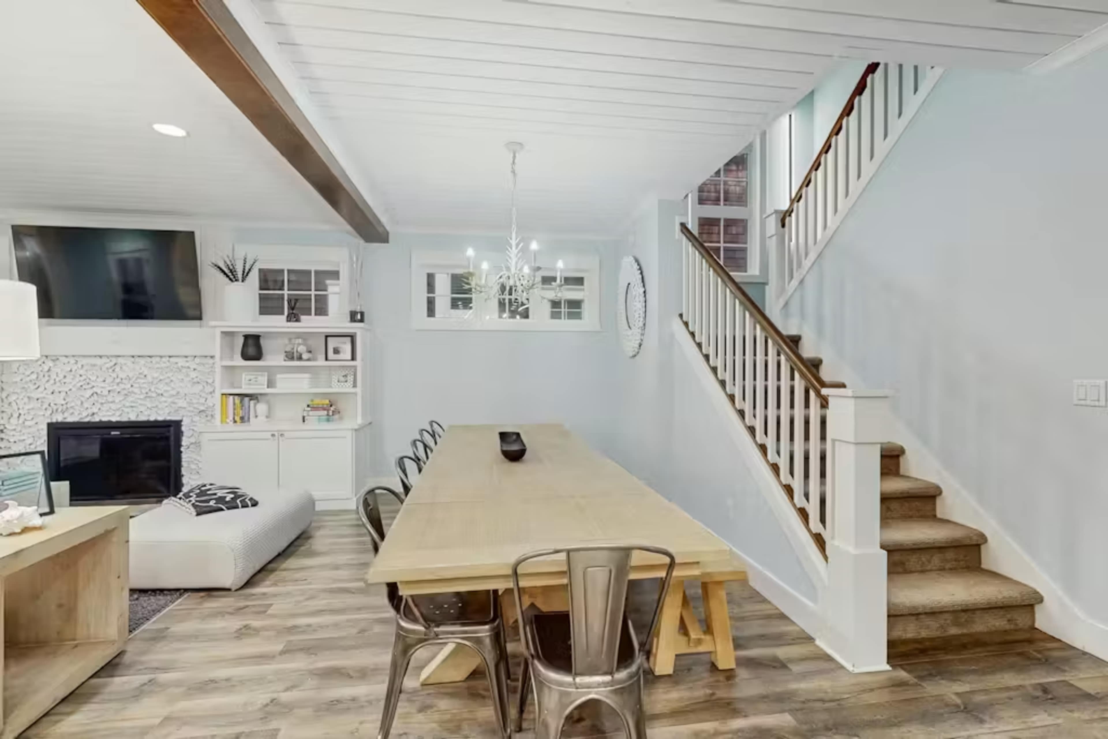 The image shows a modern dining area with a long wooden table, metal chairs, and a staircase in the background, adjacent to a living room featuring a TV and a cozy seating arrangement.