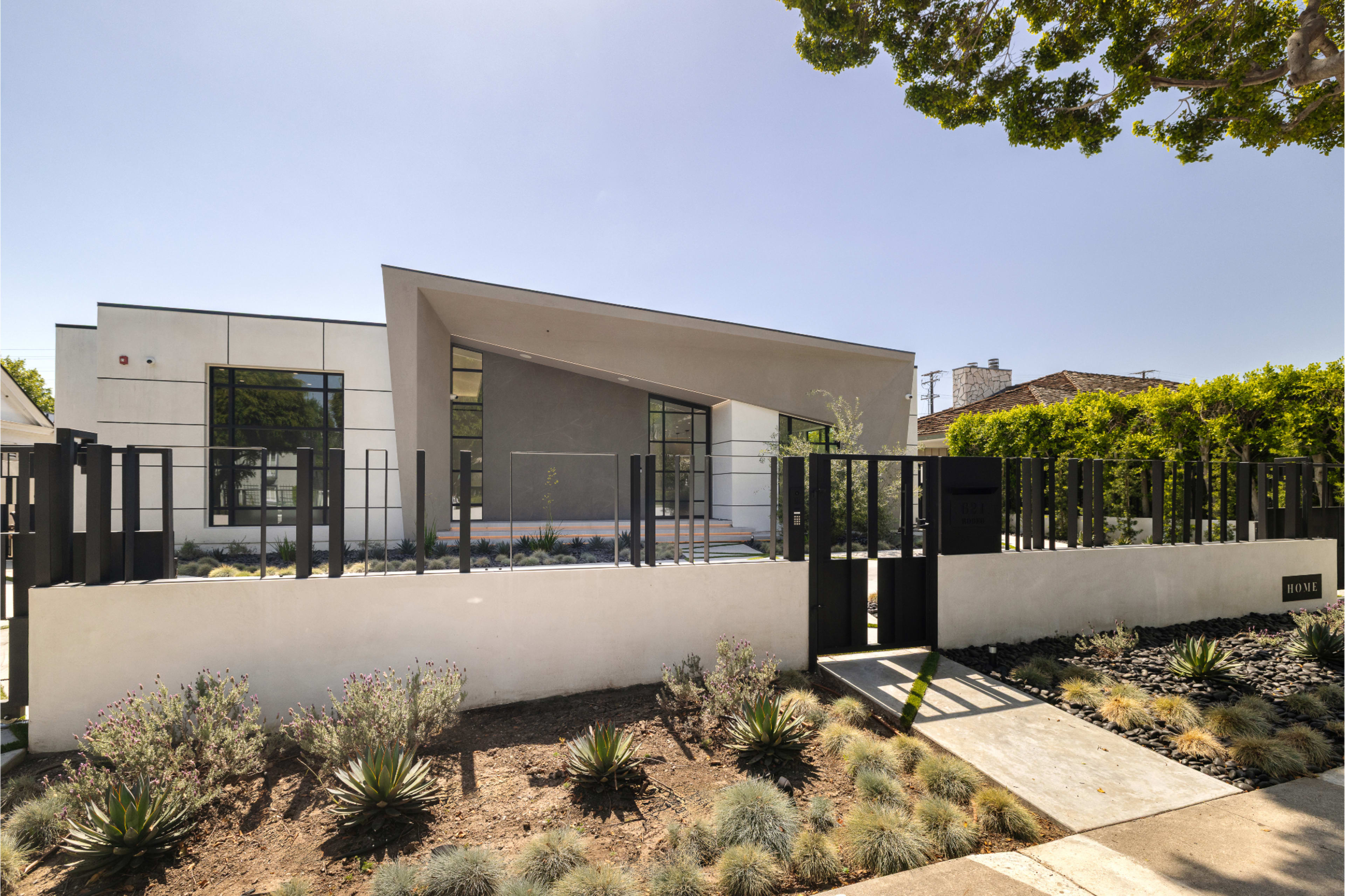 A modern house with a distinctive angled roof and a landscaped front yard is enclosed by a black fence and gate.