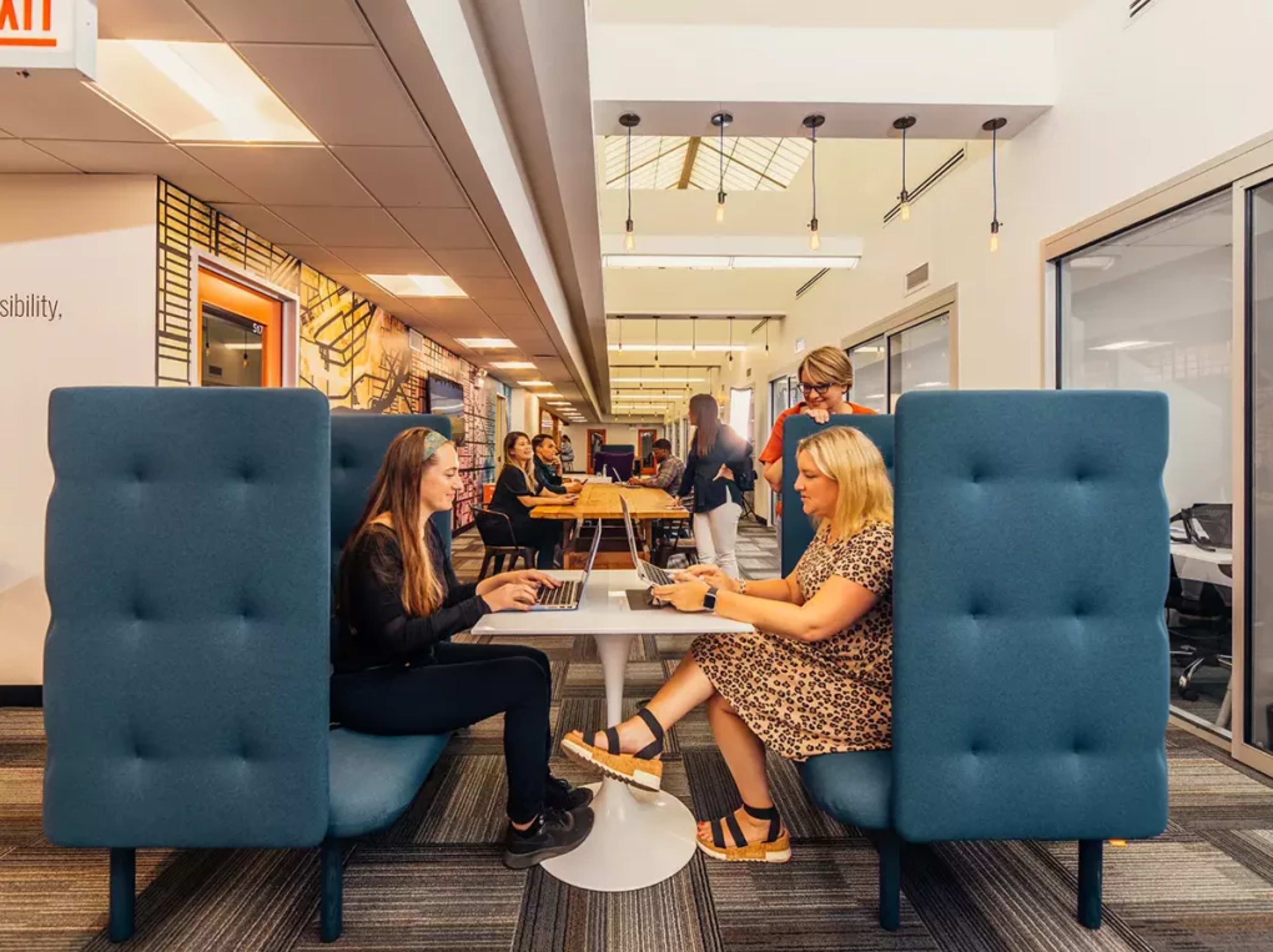 Two women are seated at a circular table in a modern office space, working on laptops while surrounded by other individuals engaged in various activities.