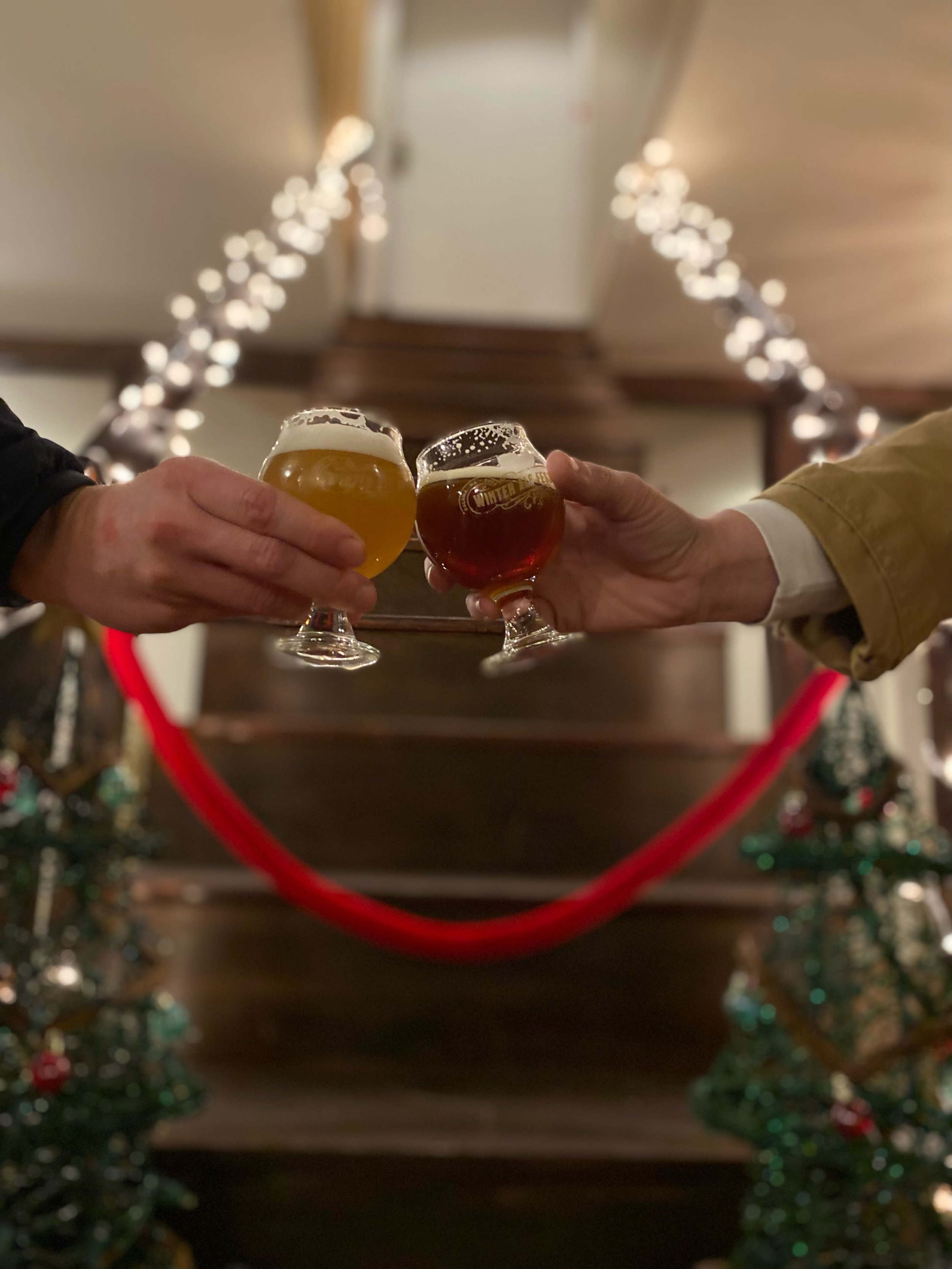 Two hands hold glasses of beer while standing on a staircase decorated with string lights and Christmas trees.
