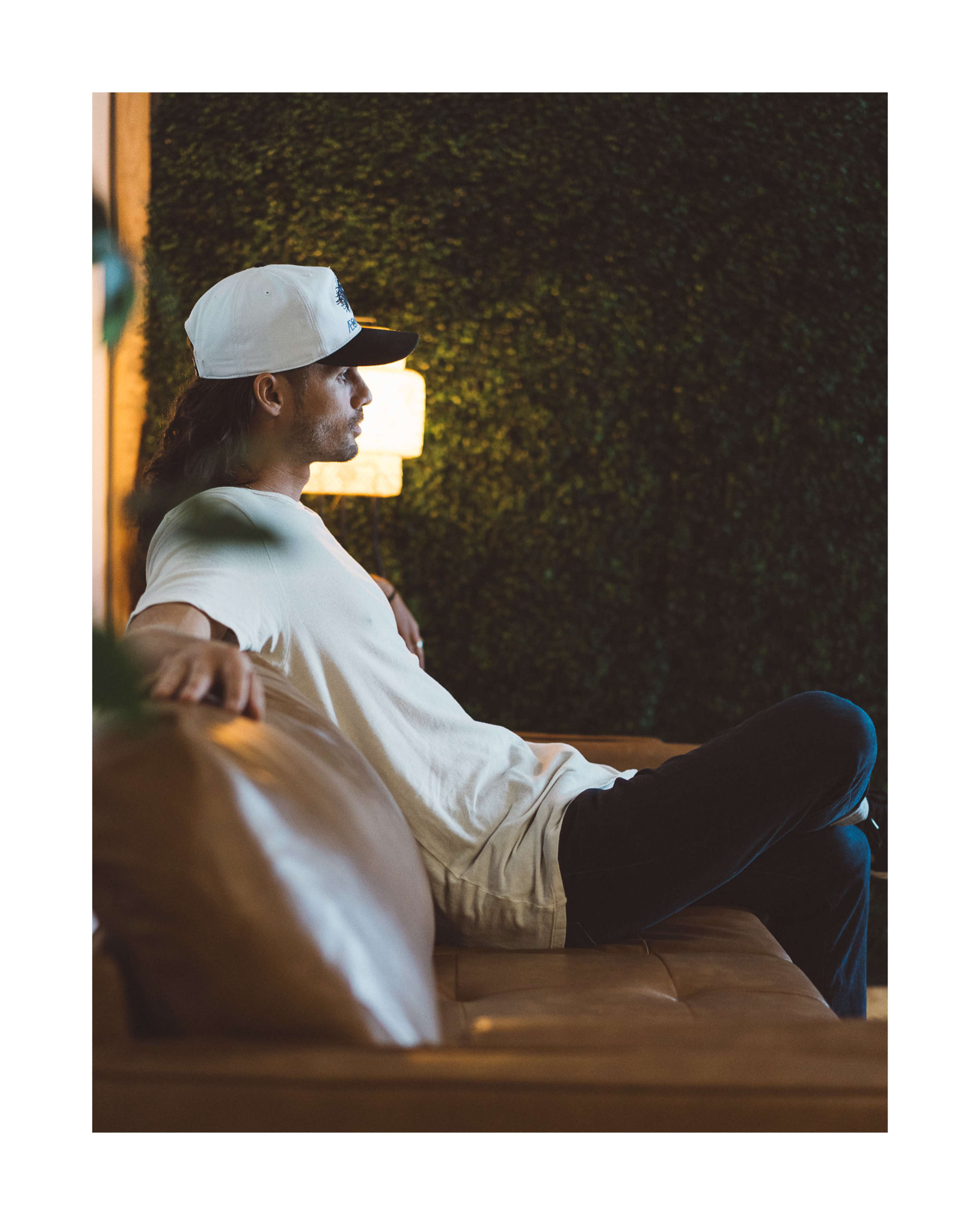 A person wearing a white shirt and a cap sits on a brown couch against a green textured wall.
