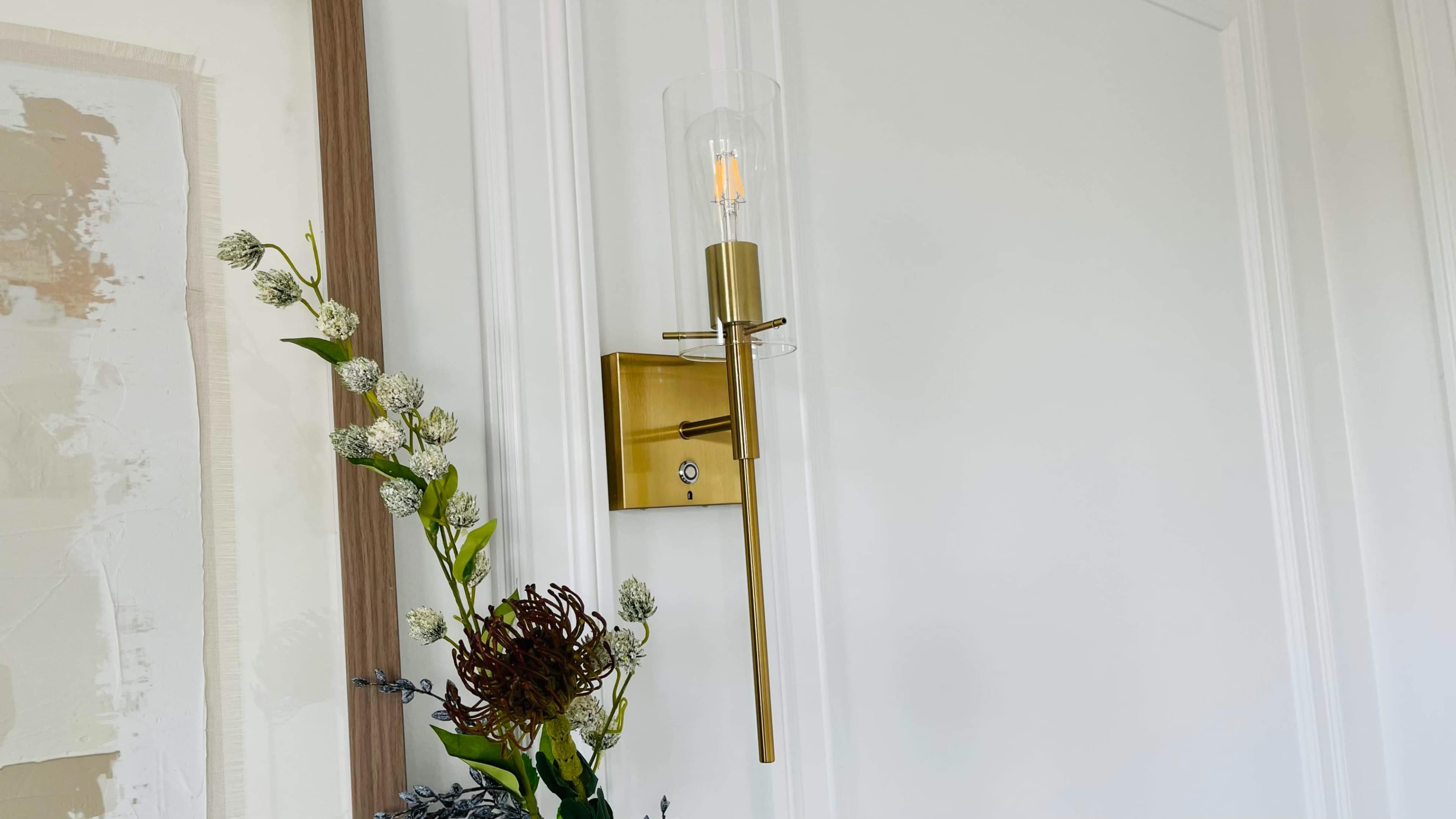 The image shows a modern wall sconce with a clear glass shade and a gold finish, mounted beside a floral arrangement.
