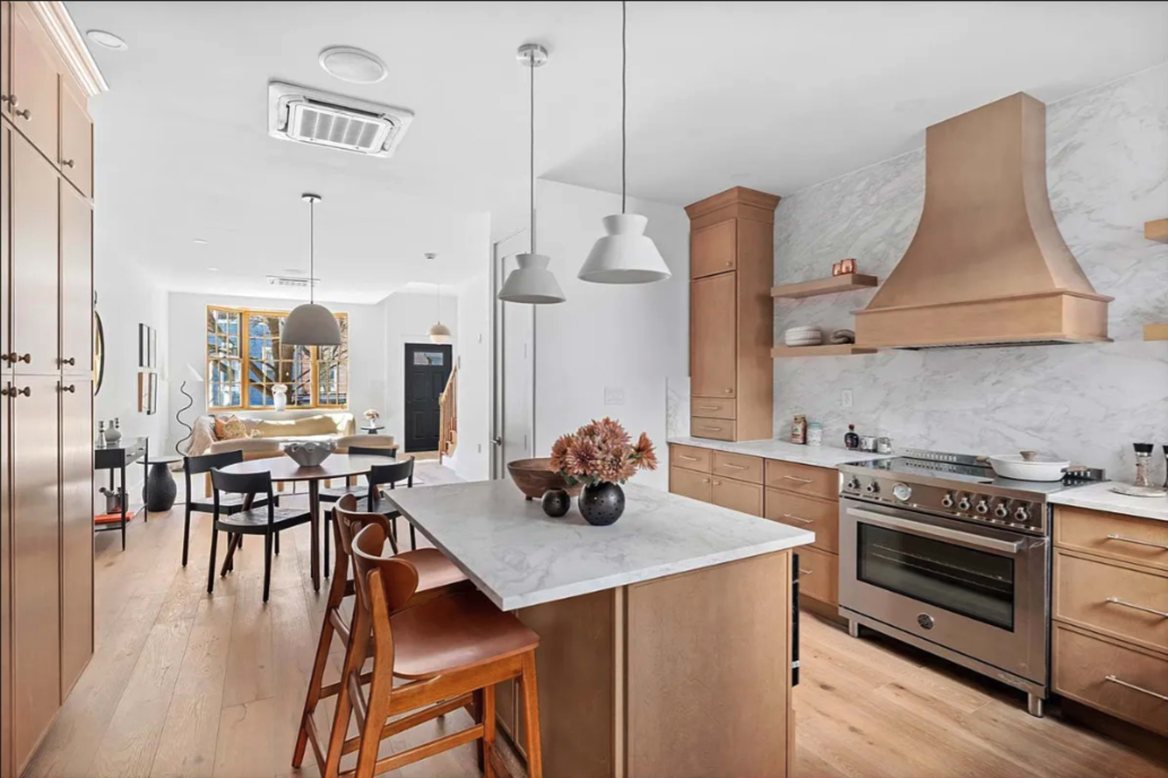 The image shows a modern kitchen featuring a large marble island, stainless steel appliances, and wooden cabinetry, with a dining area visible in the background.