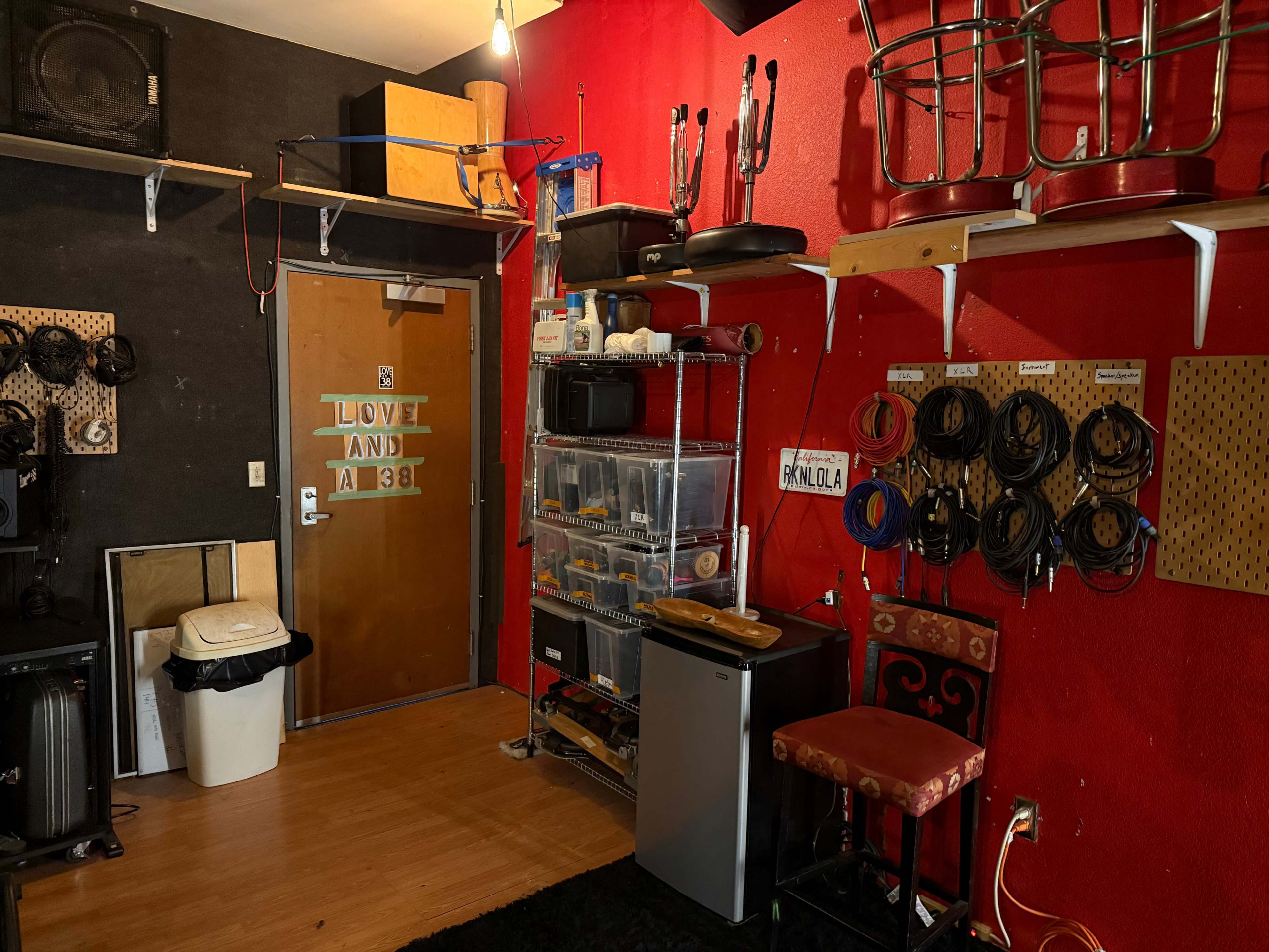 The image shows a room with a red wall, displaying shelves filled with various equipment, a mini fridge, and an entrance door with the words "LOVE AND A38" on it.