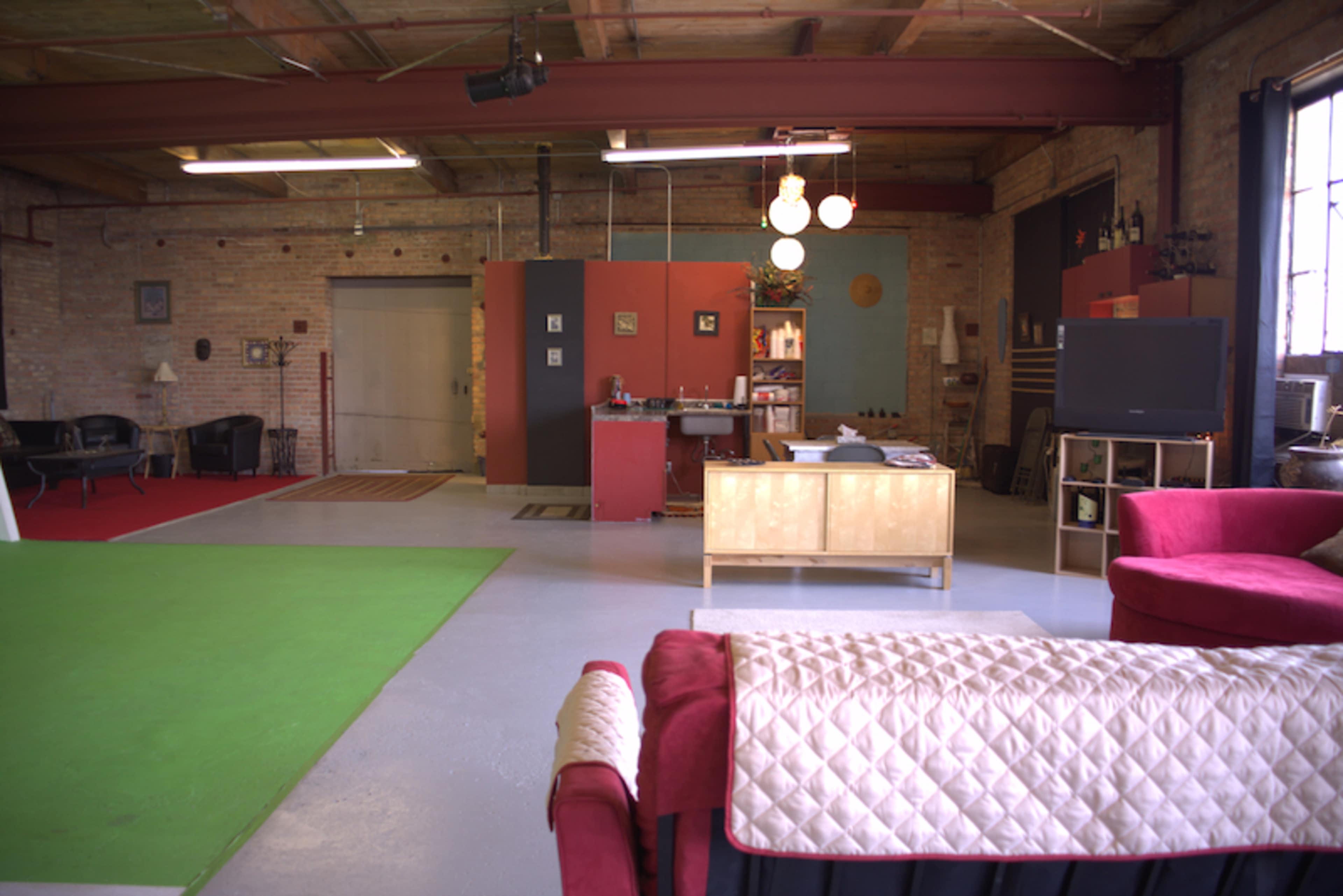 A spacious industrial loft with a red and green area rug, a sofa, a small kitchen area, and various furniture items.