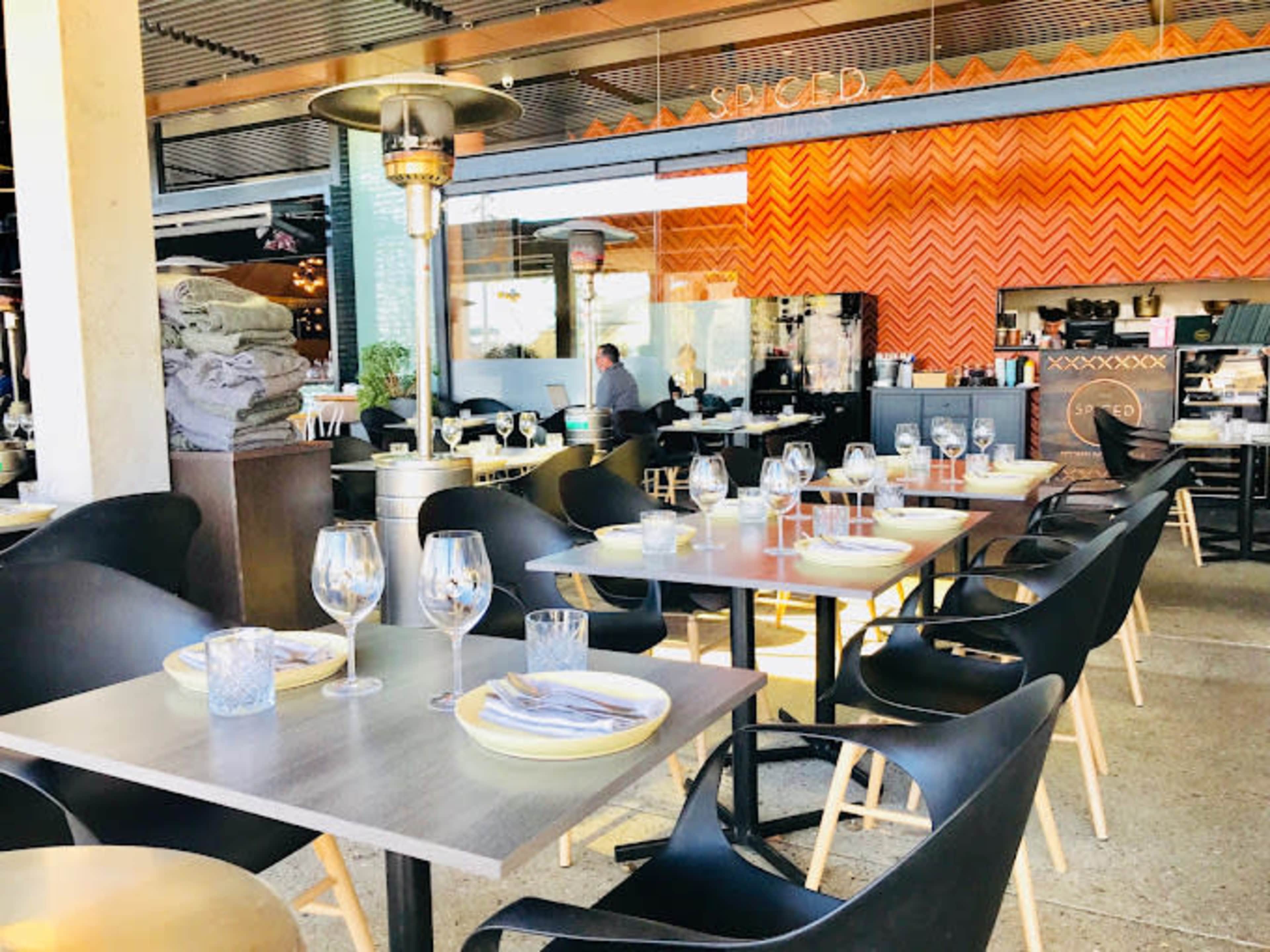 The image shows an outdoor dining area of a restaurant with tables set for patrons, featuring chairs and heaters, alongside a contemporary interior visible in the background.