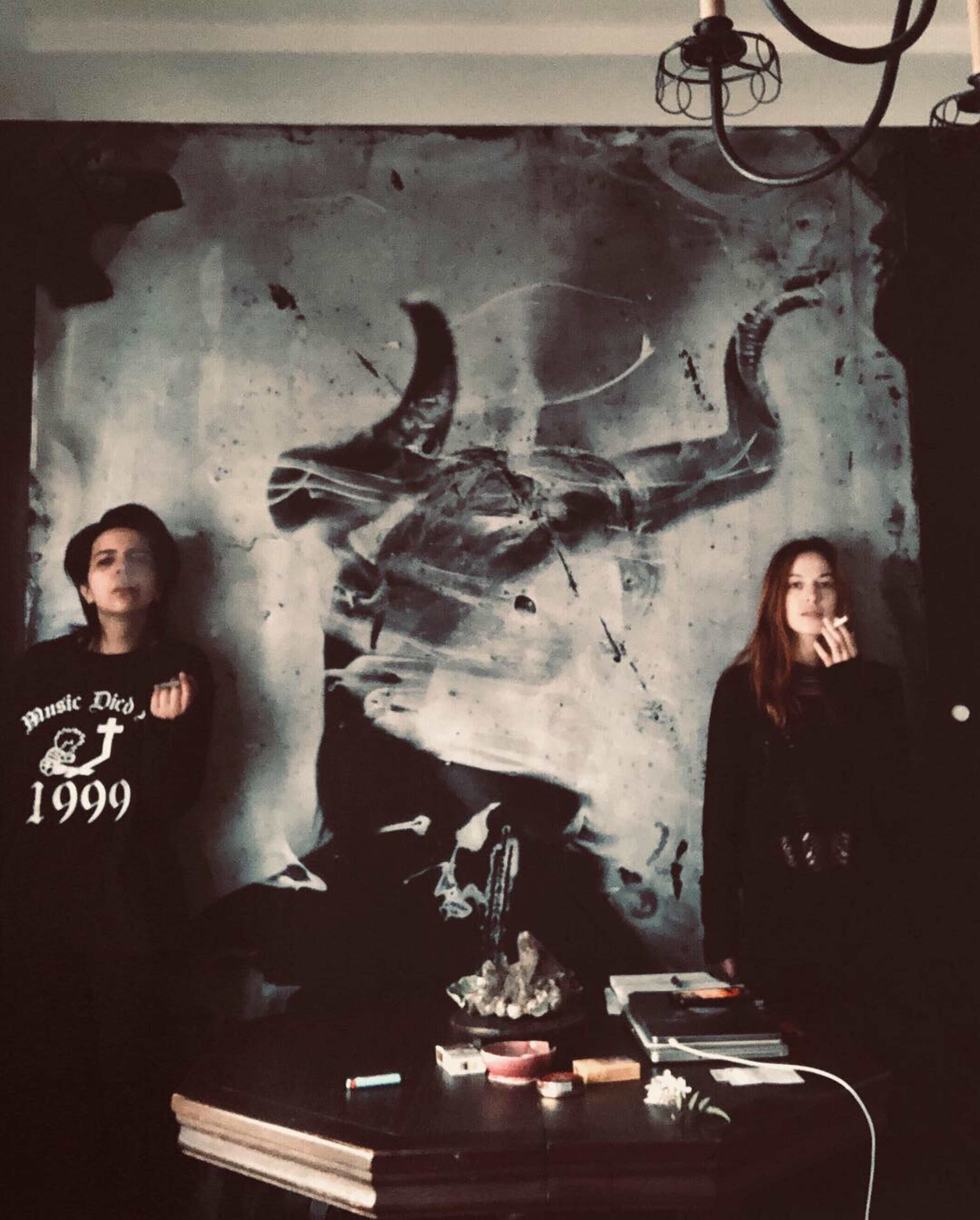 Two individuals stand beside a large, dark wall mural depicting a bull skull, with a table in front holding various items.