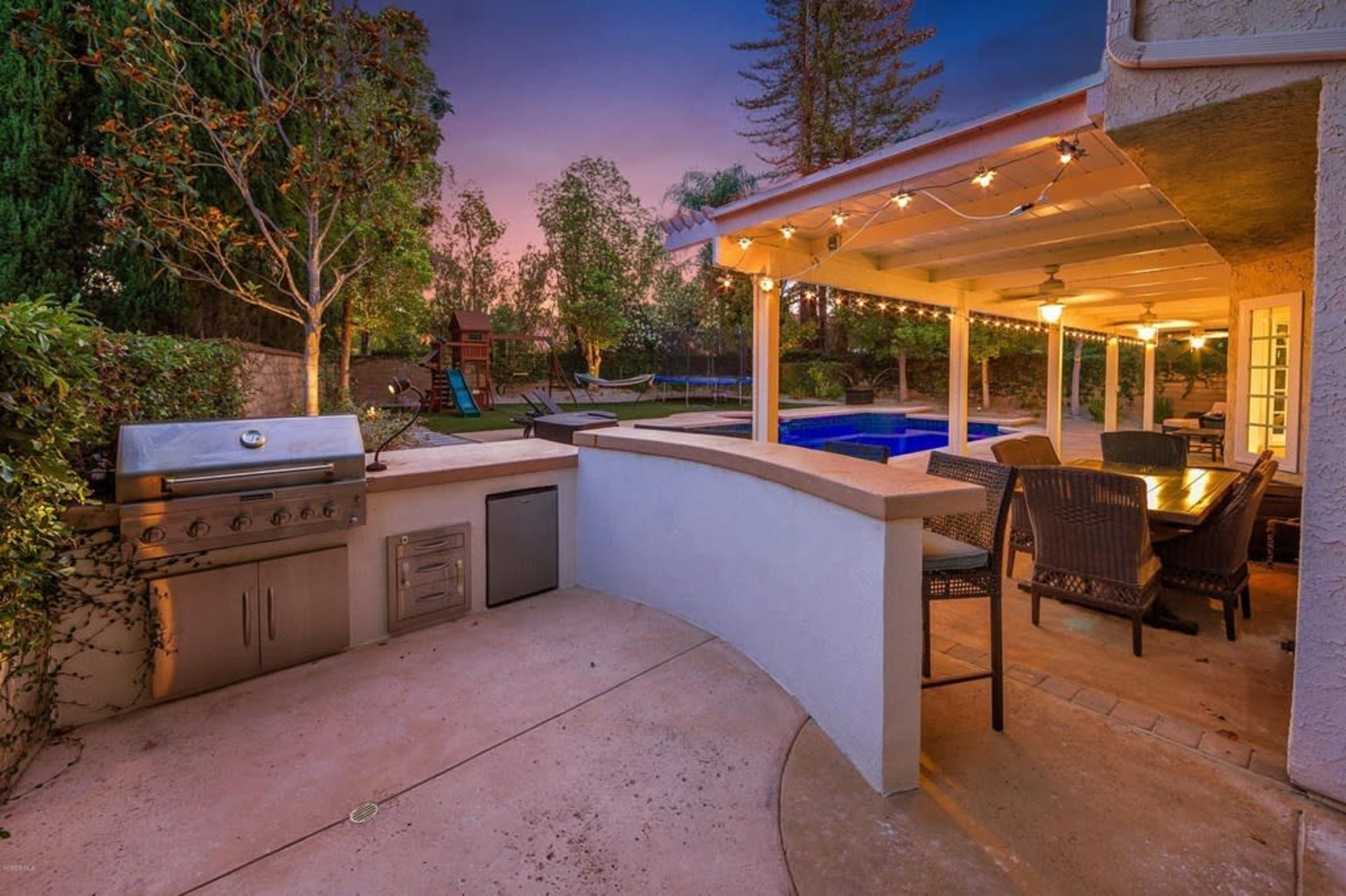 The image shows an outdoor patio area with a barbecue grill, dining table, and a pool, surrounded by trees and decorative string lights.