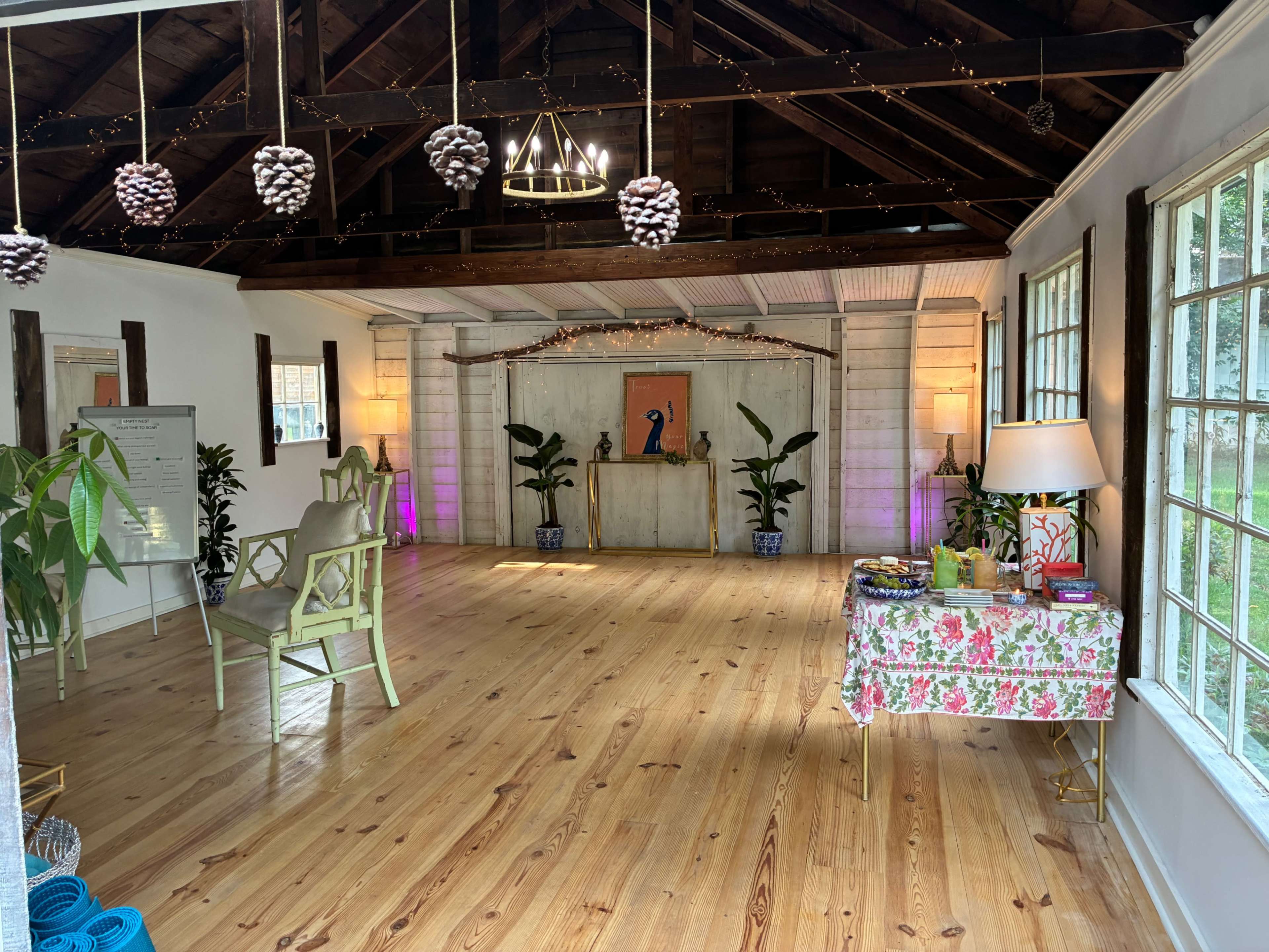 The interior of a spacious, well-lit room features wooden flooring, decorated with potted plants, a floral tablecloth, and a simple art display in the background.