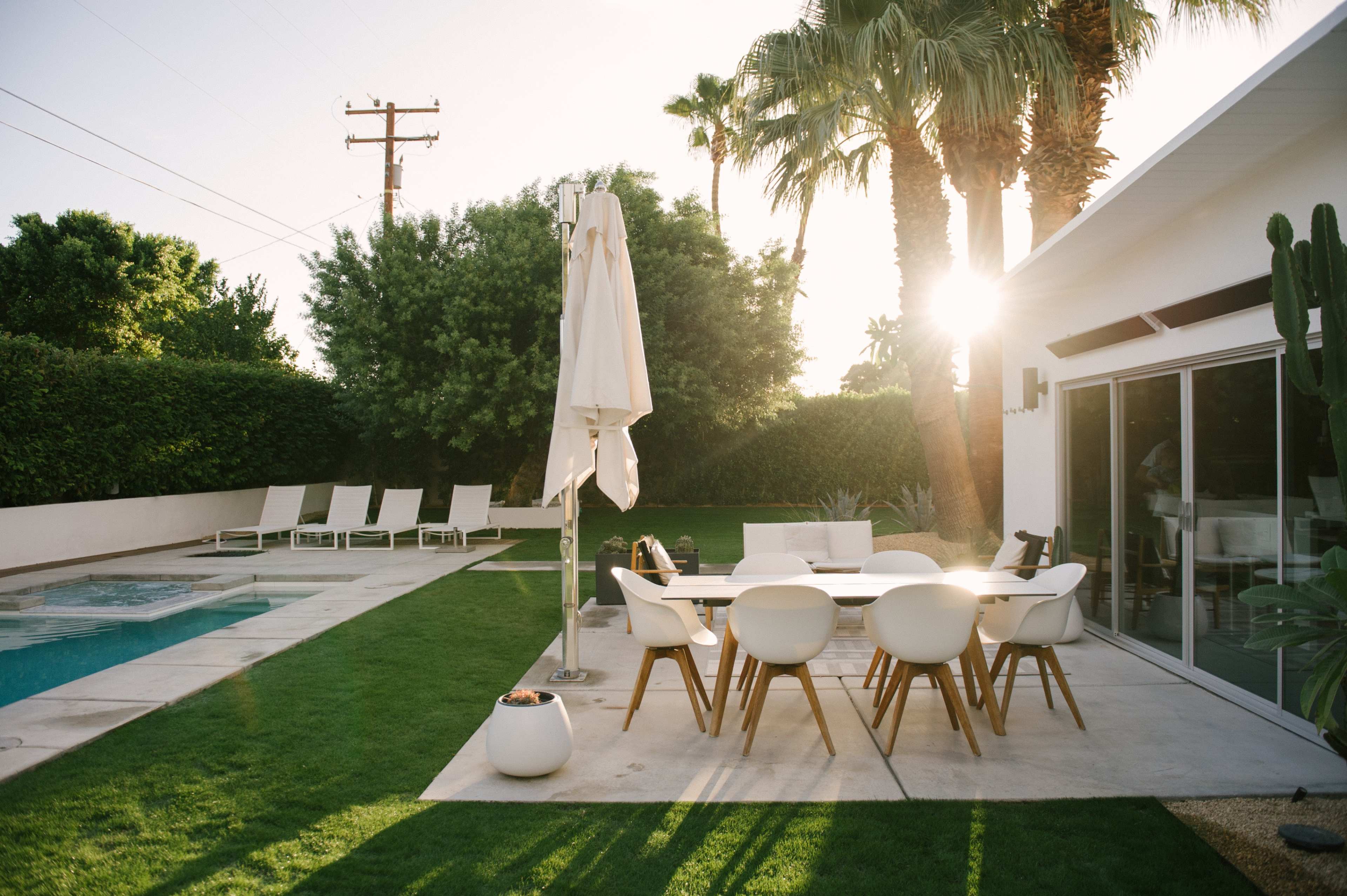 A modern outdoor dining area is situated next to a swimming pool, surrounded by palm trees and lounge chairs, with sunlight filtering through the foliage.