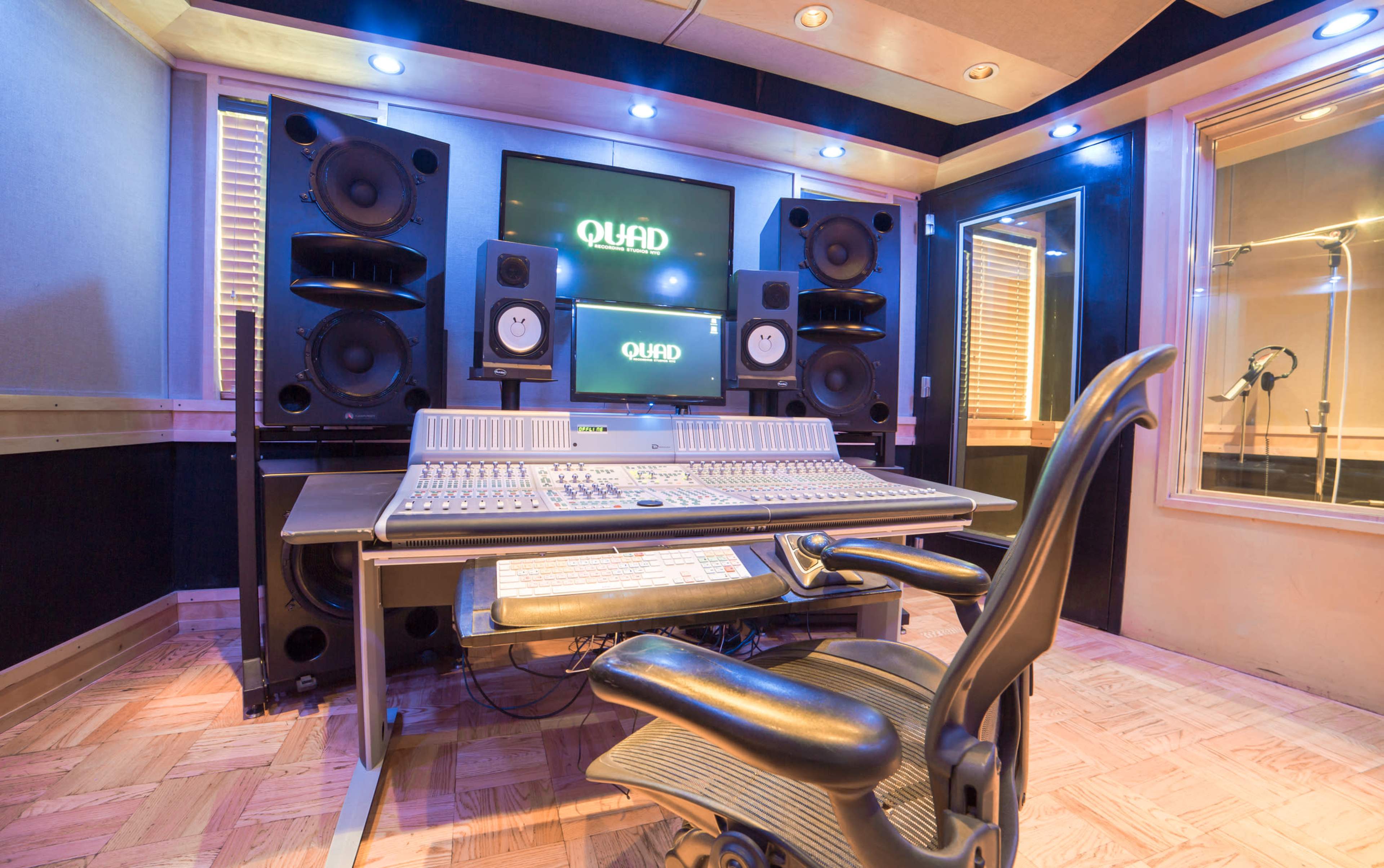 The image shows a music studio featuring a mixing console, large speakers, a monitor display, and a recording booth in the background.