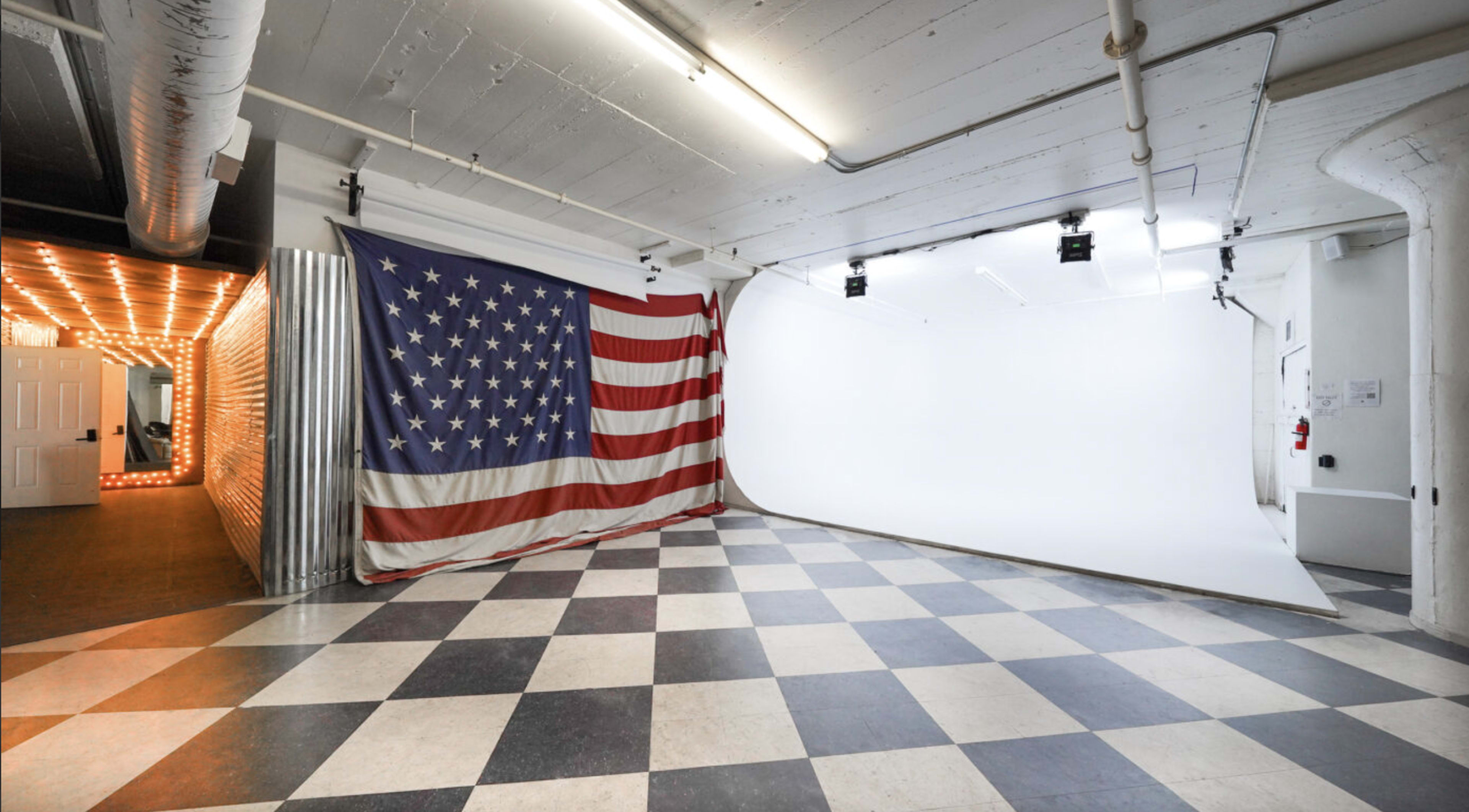 A spacious studio with a large American flag on one wall and a white backdrop on the opposite side, all set against a checkered floor.
