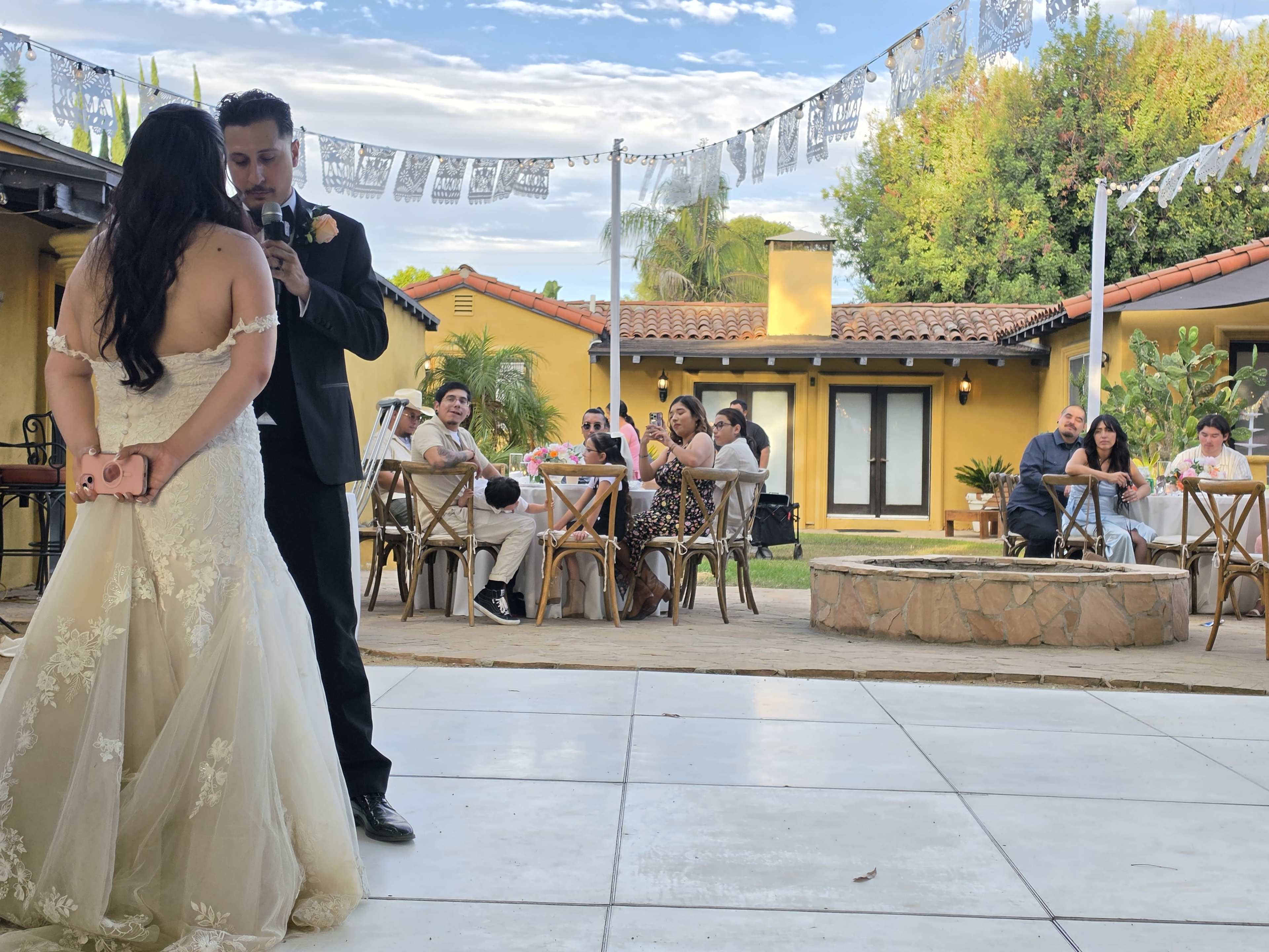 A man in a suit speaks to a woman in a wedding dress while guests sit at tables in a courtyard decorated with paper banners.