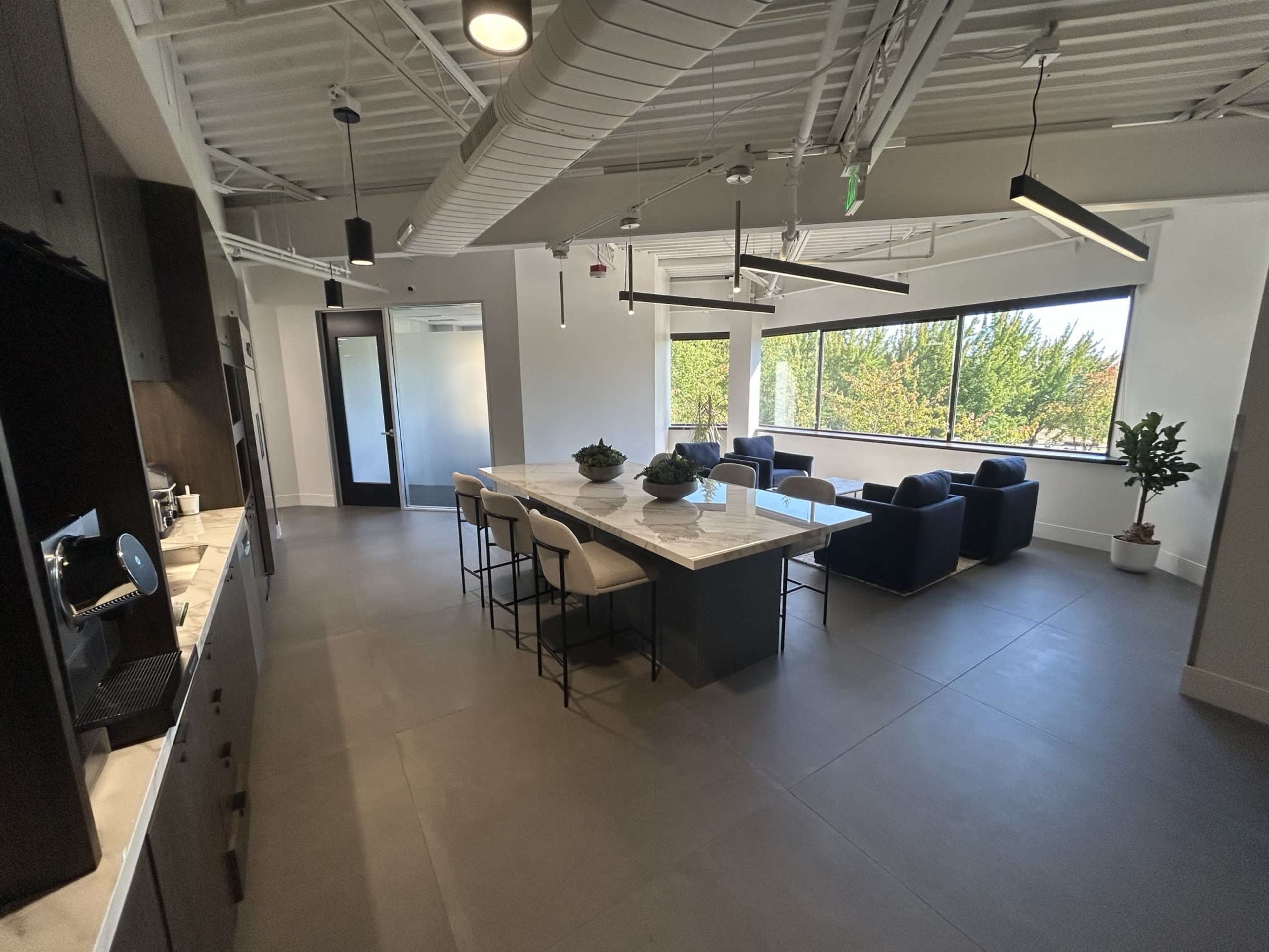 The image shows a modern office space with a kitchen area, a large central island with seating, and a lounge with blue chairs next to a window.