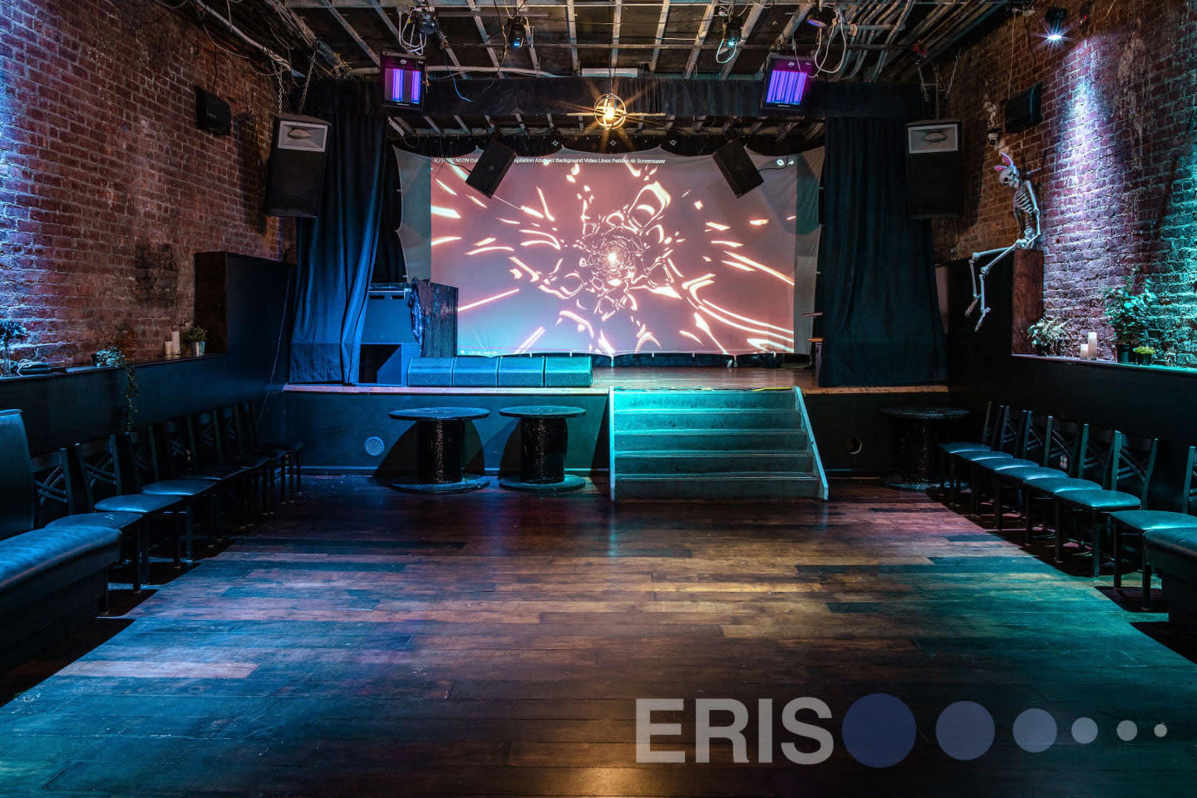 An empty music venue with a stage displaying visual projections, surrounded by seating areas and dim lighting.