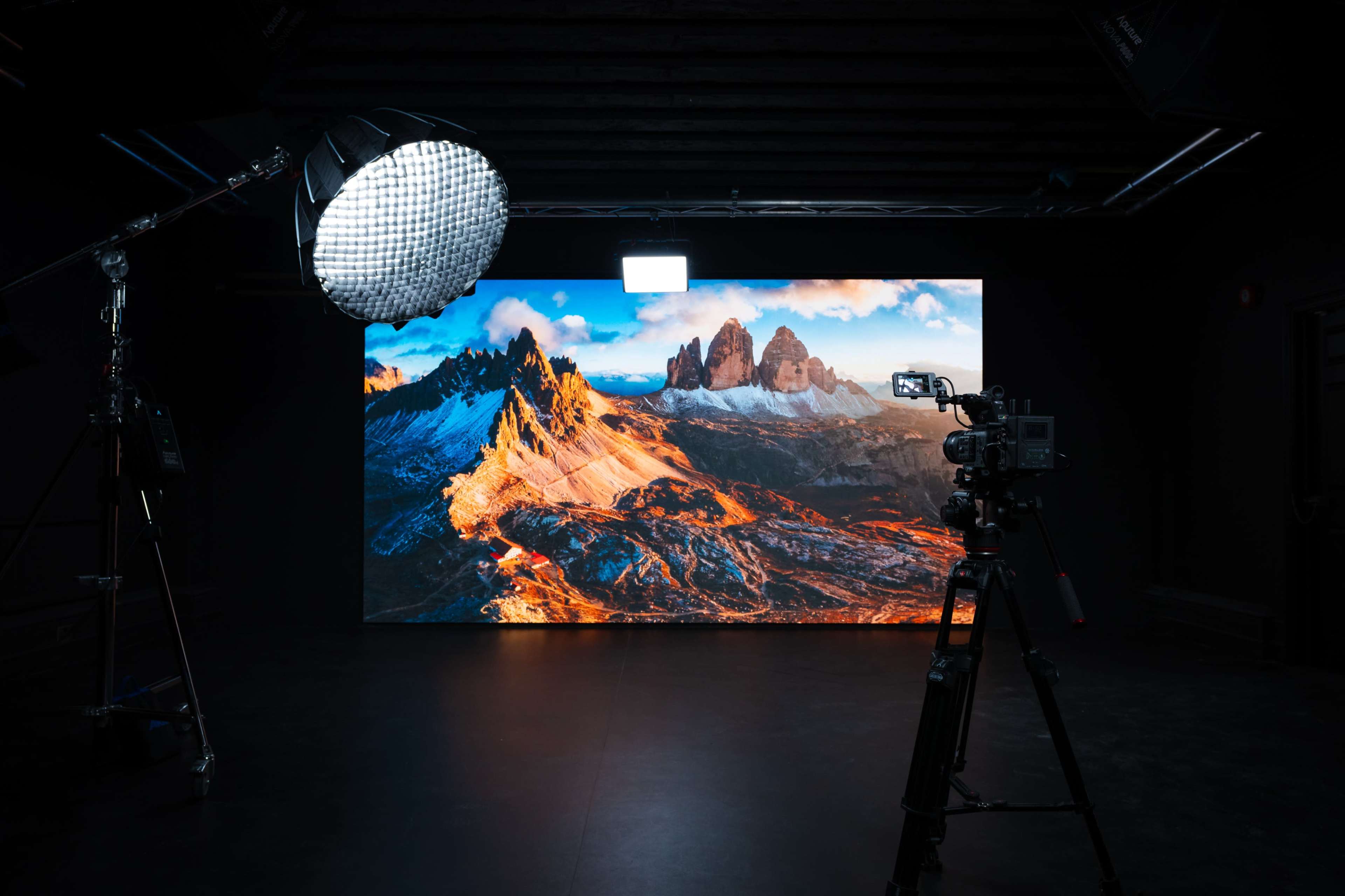 The image shows a large, vibrant screen displaying a mountainous landscape, with a camera and lighting equipment positioned in a dark studio setting.
