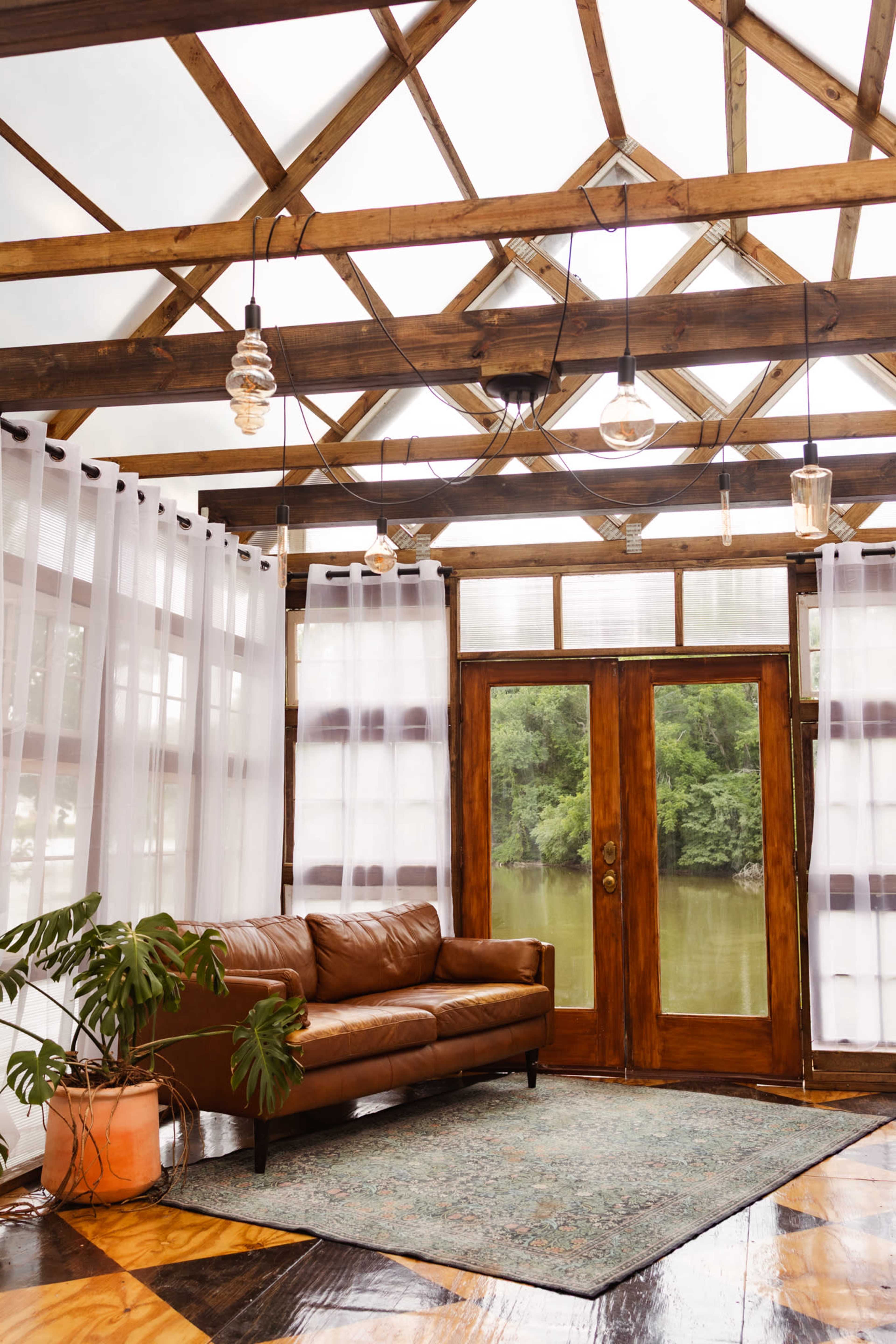 The interior features a brown leather sofa facing large double doors that open to a view of a river, with a geometric wooden ceiling and sheer white curtains.