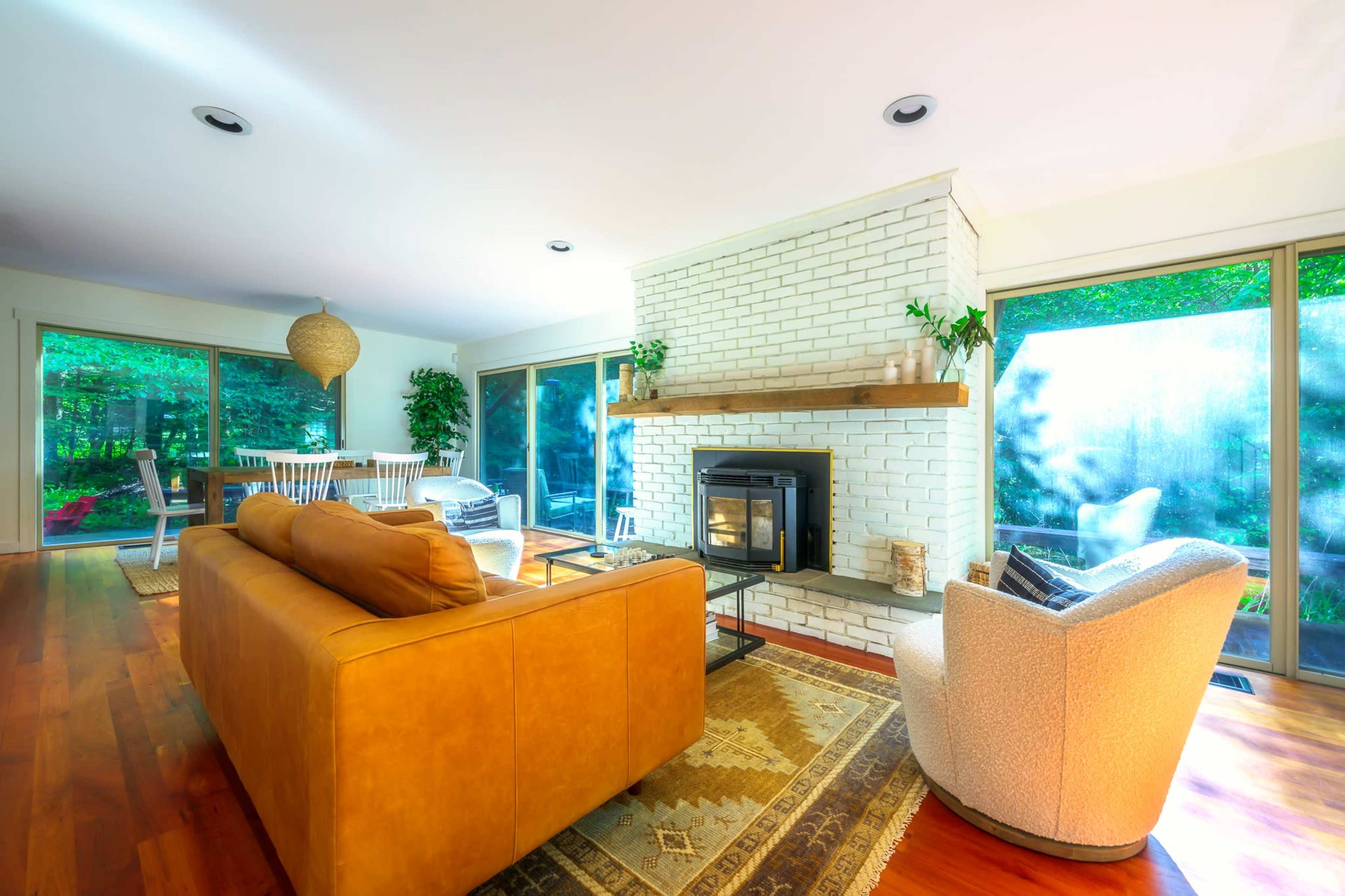 A modern living room with a brick fireplace, a tan couch, a white armchair, and large windows overlooking a garden.