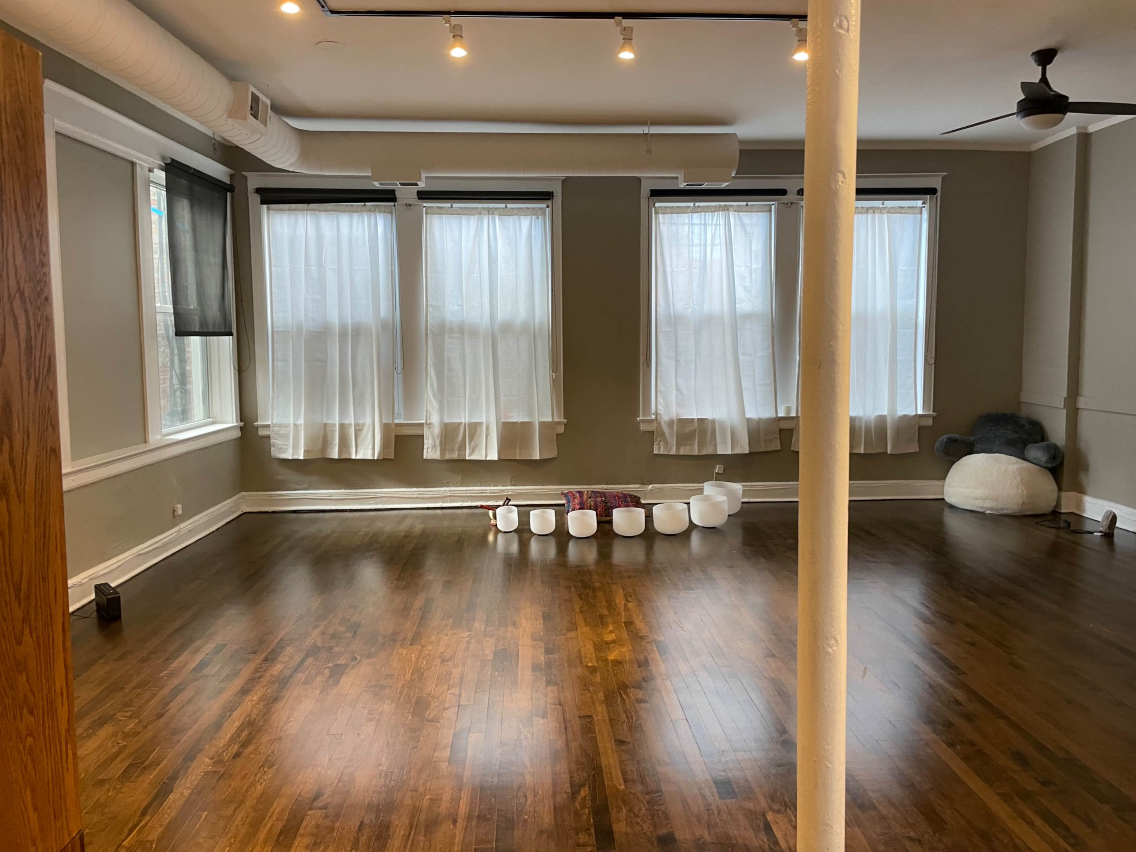 The image shows a spacious room with wooden floors, three windows covered by sheer curtains, and a row of white bowls arranged on the floor.