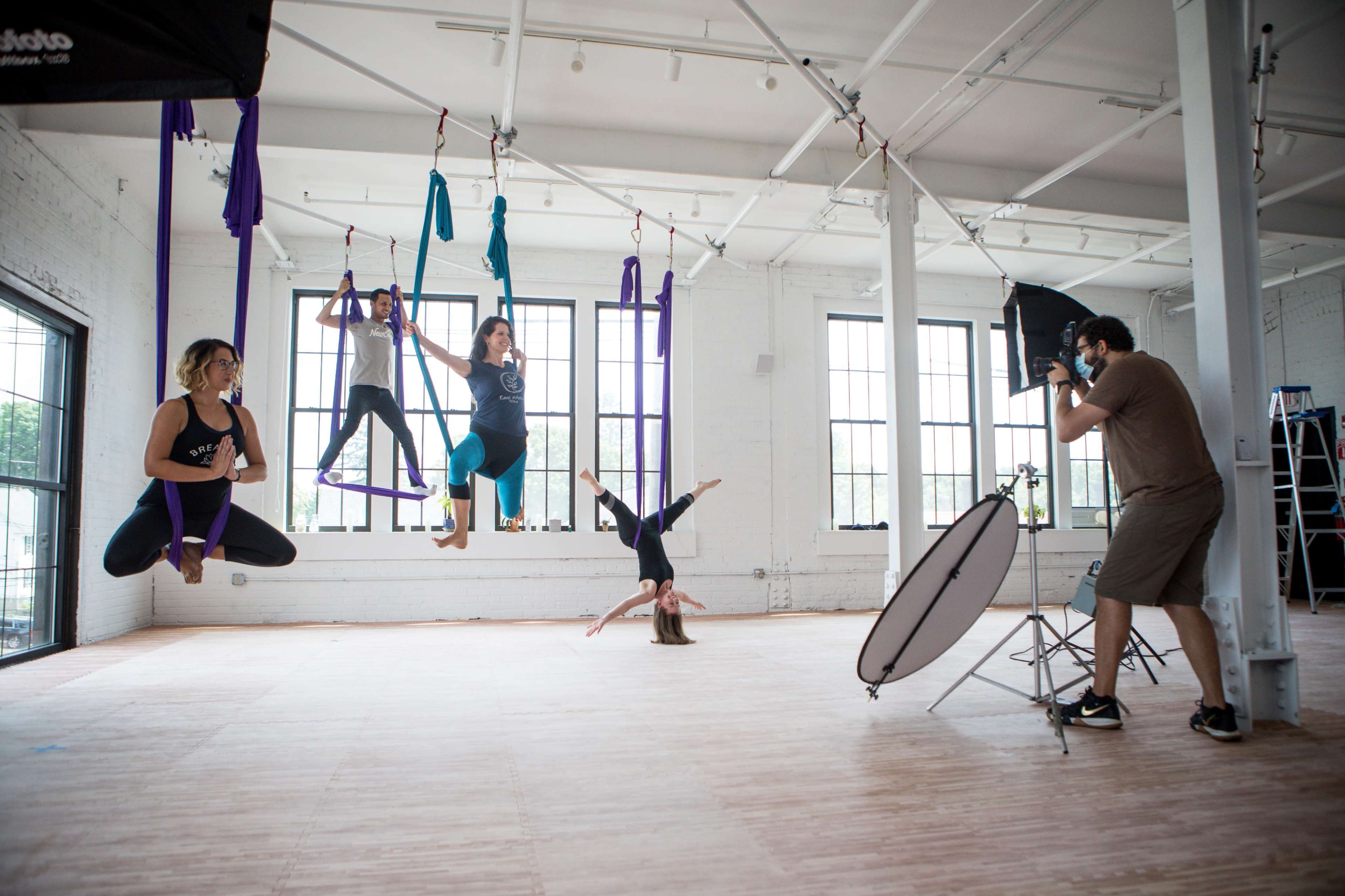 A group of women performs aerial silk gymnastic poses in a spacious studio while a photographer captures their movements.
