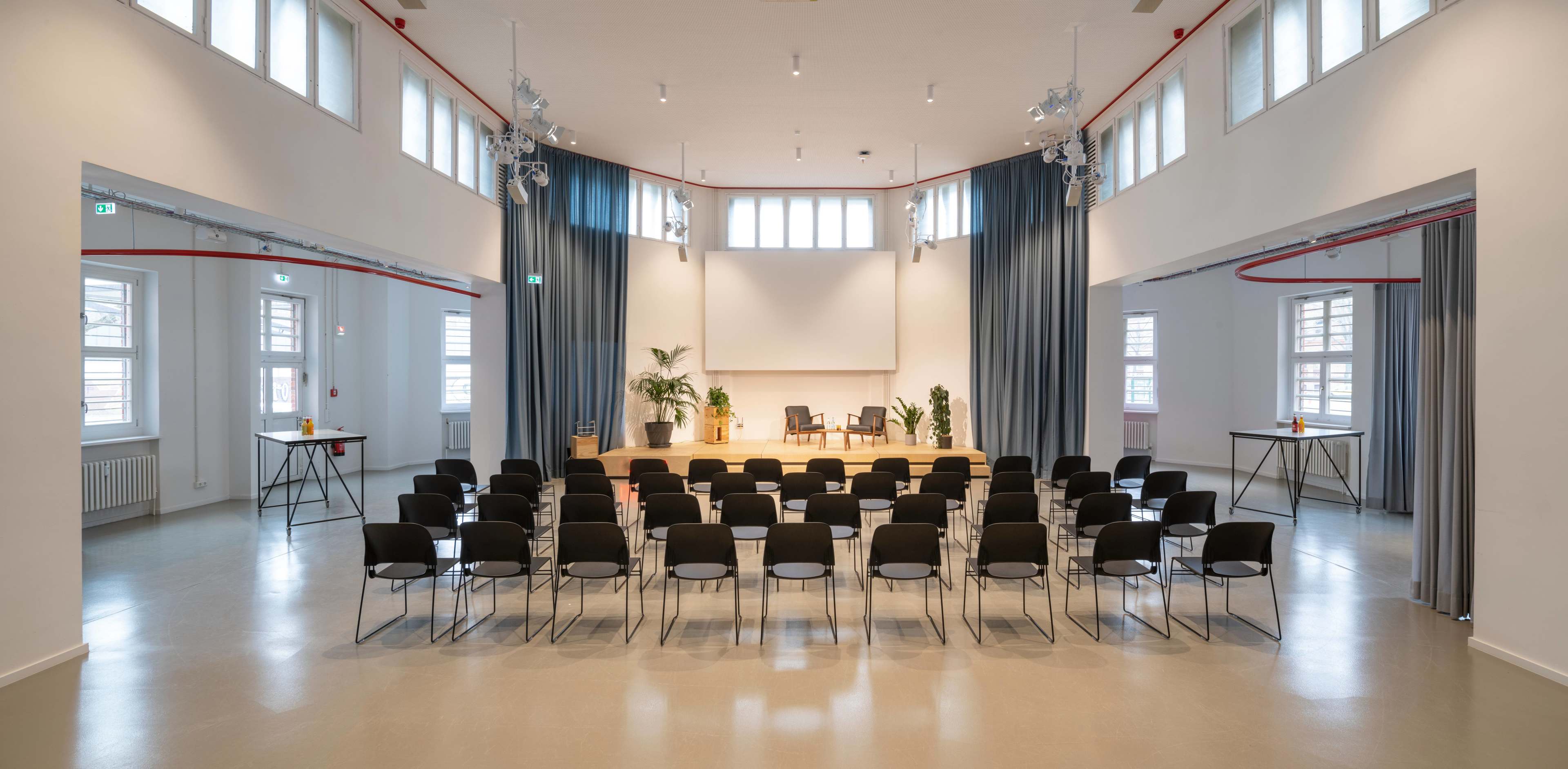 The image shows a spacious indoor venue set up with rows of black chairs facing a wooden stage that features two chairs and a large screen, with curtains and plants decorating the surrounding area.