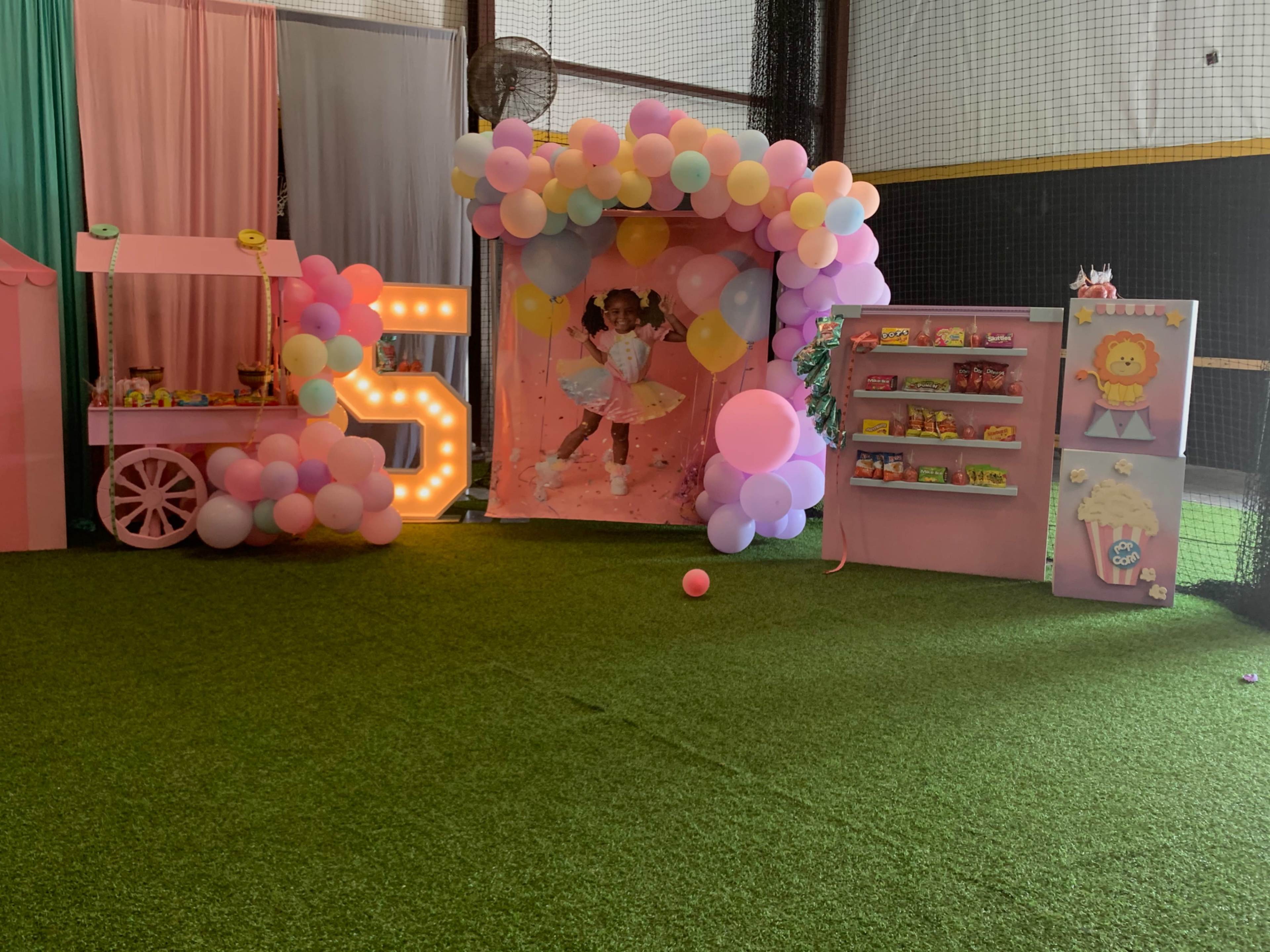 The image shows a brightly decorated indoor party area featuring a pink backdrop with balloons, a cart with snacks, and colorful shelves displaying treats.