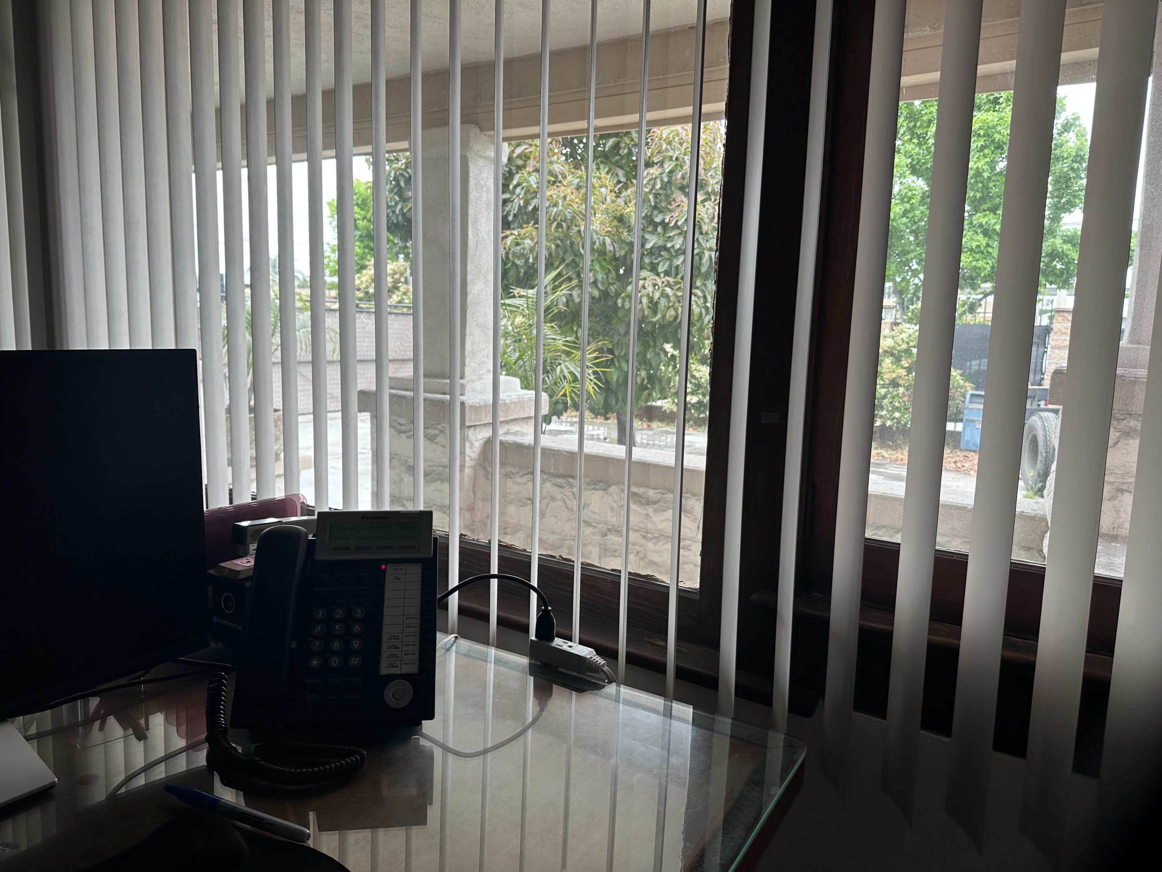 A desk with a telephone and computer is visible beside a window covered with vertical blinds, which reveals outdoor greenery and a stone wall.