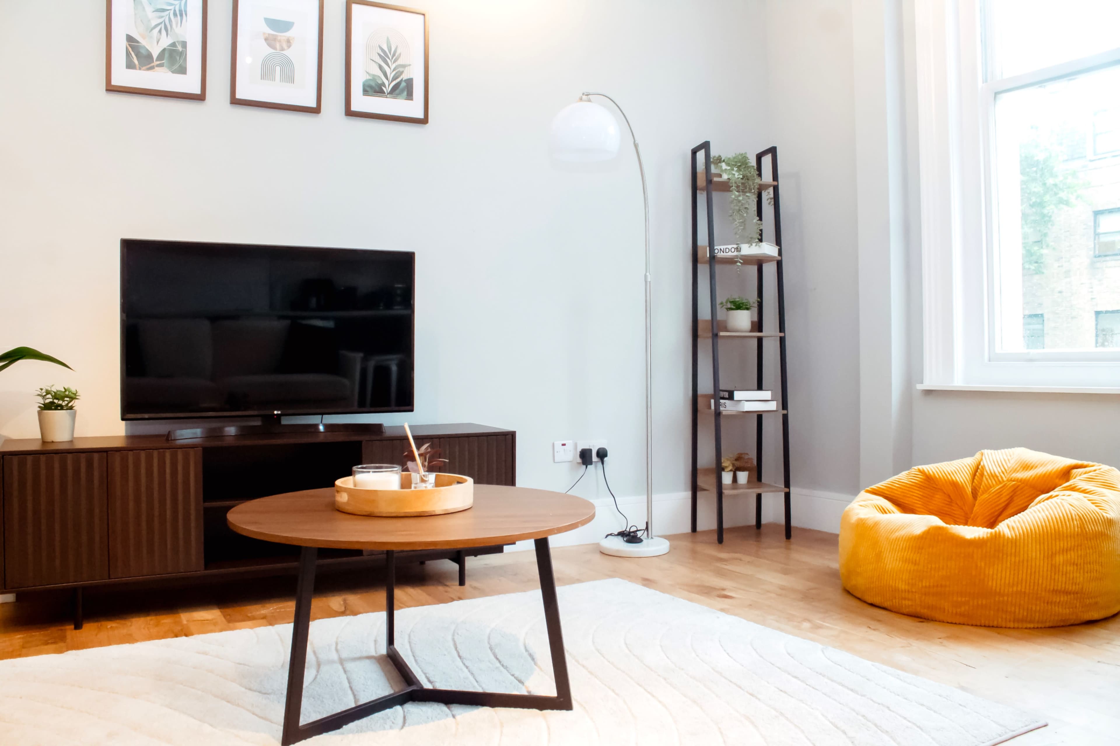 The image shows a modern living room with a television mounted on a wooden cabinet, a round coffee table, a cozy bean bag, and a tall shelf displaying decorative items.