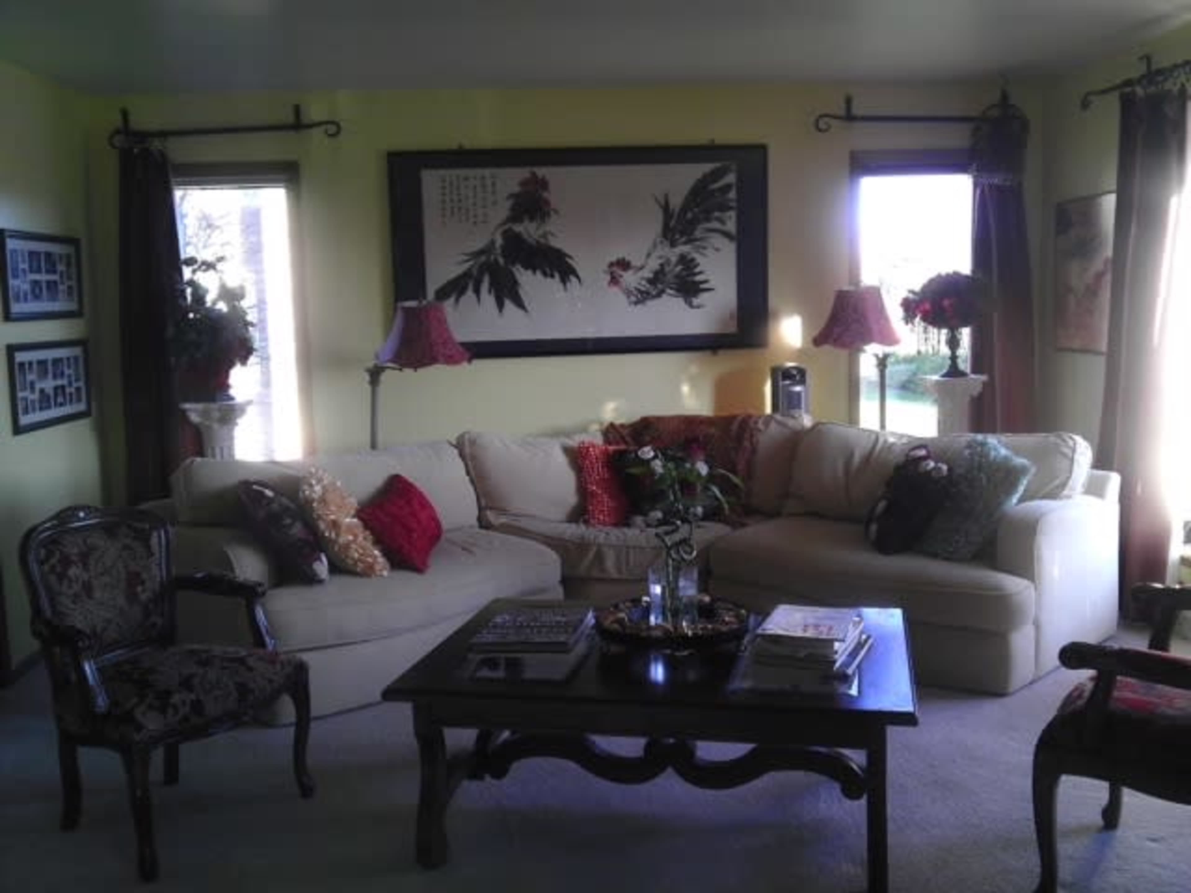 The image shows a cozy living room with a light-colored sectional sofa, a dark wooden coffee table, decorative pillows, and large windows allowing natural light to enter.