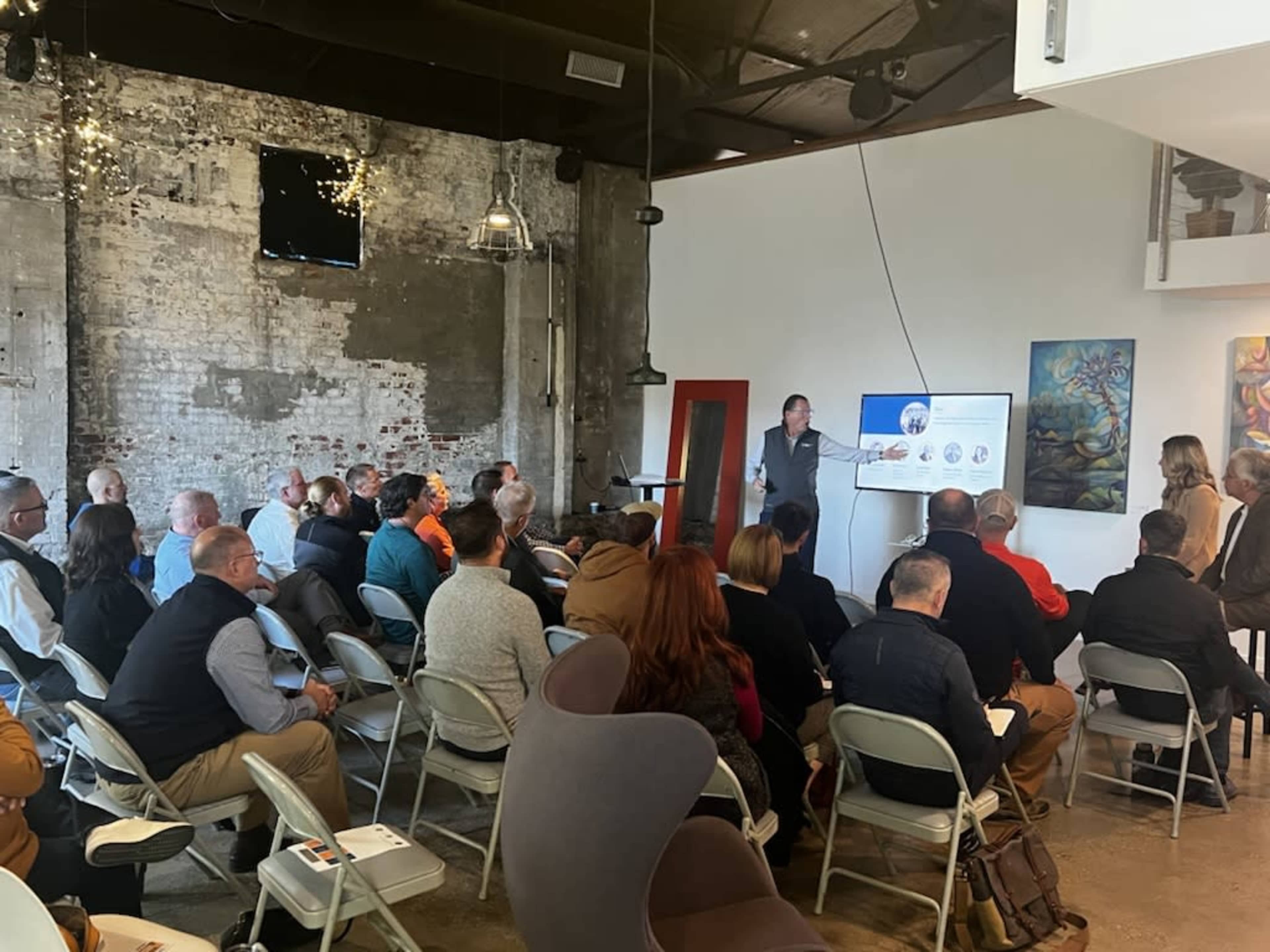 A speaker presents to an audience seated in a modern, industrial-style venue with exposed brick walls and artwork on display.