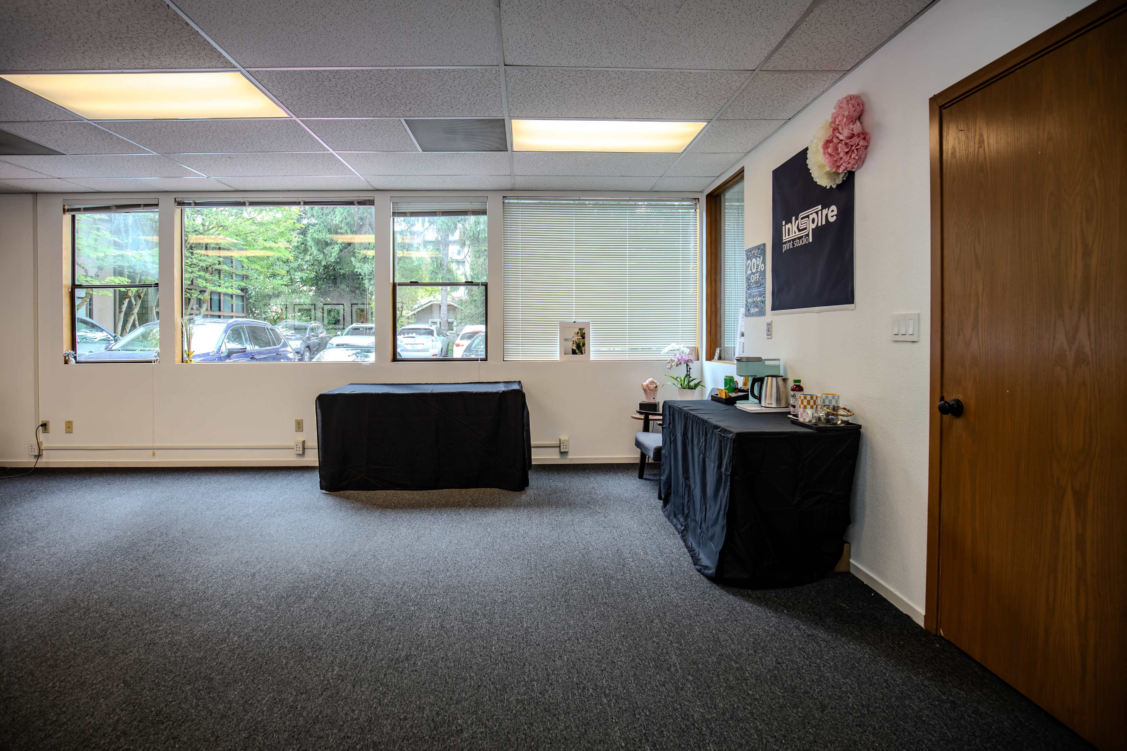 The image shows an empty room with two tables covered by black cloths, large windows allowing natural light, and a door on one side.